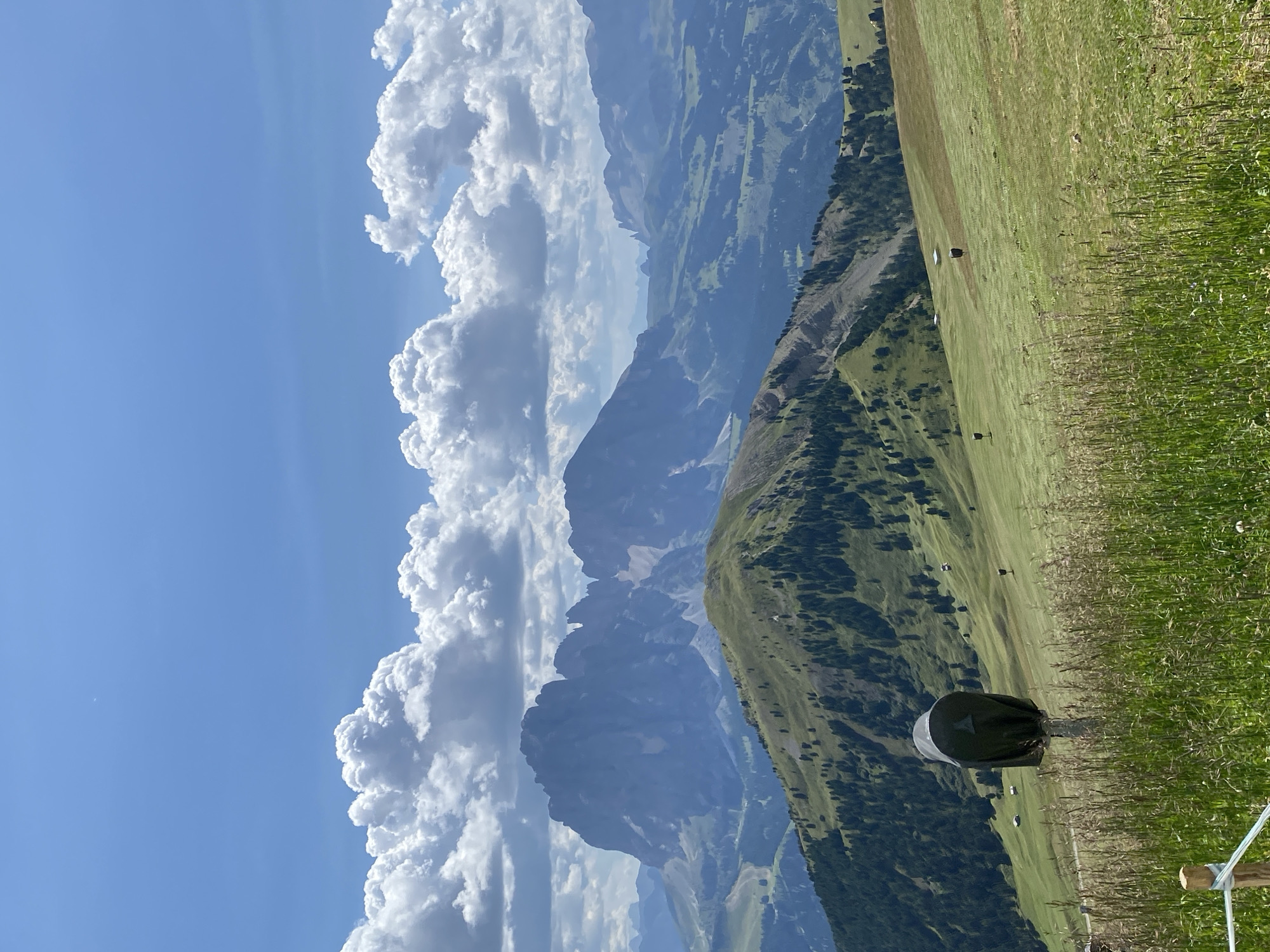 This is a landscape photograph taken in Santa Cristina Gherdëina, Italy. The scene is a mountainous region with rolling green hills covered in grass and scattered trees. In the foreground, tall grass and a wooden post with a dark, covered object are visible. Further up the hillside, several small, dark objects resembling covered equipment or markers are dotted across the landscape. In the mid-ground, a prominent green hill with a ridge line is covered with trees. In the background, massive, jagged mountain peaks dominate the horizon, some with patches of snow. The sky is a vibrant blue with large, fluffy white clouds. The image depicts a natural, outdoor environment, likely a mountain resort or hiking area, under a bright, partly cloudy sky. There are no people or animals visible in the frame.