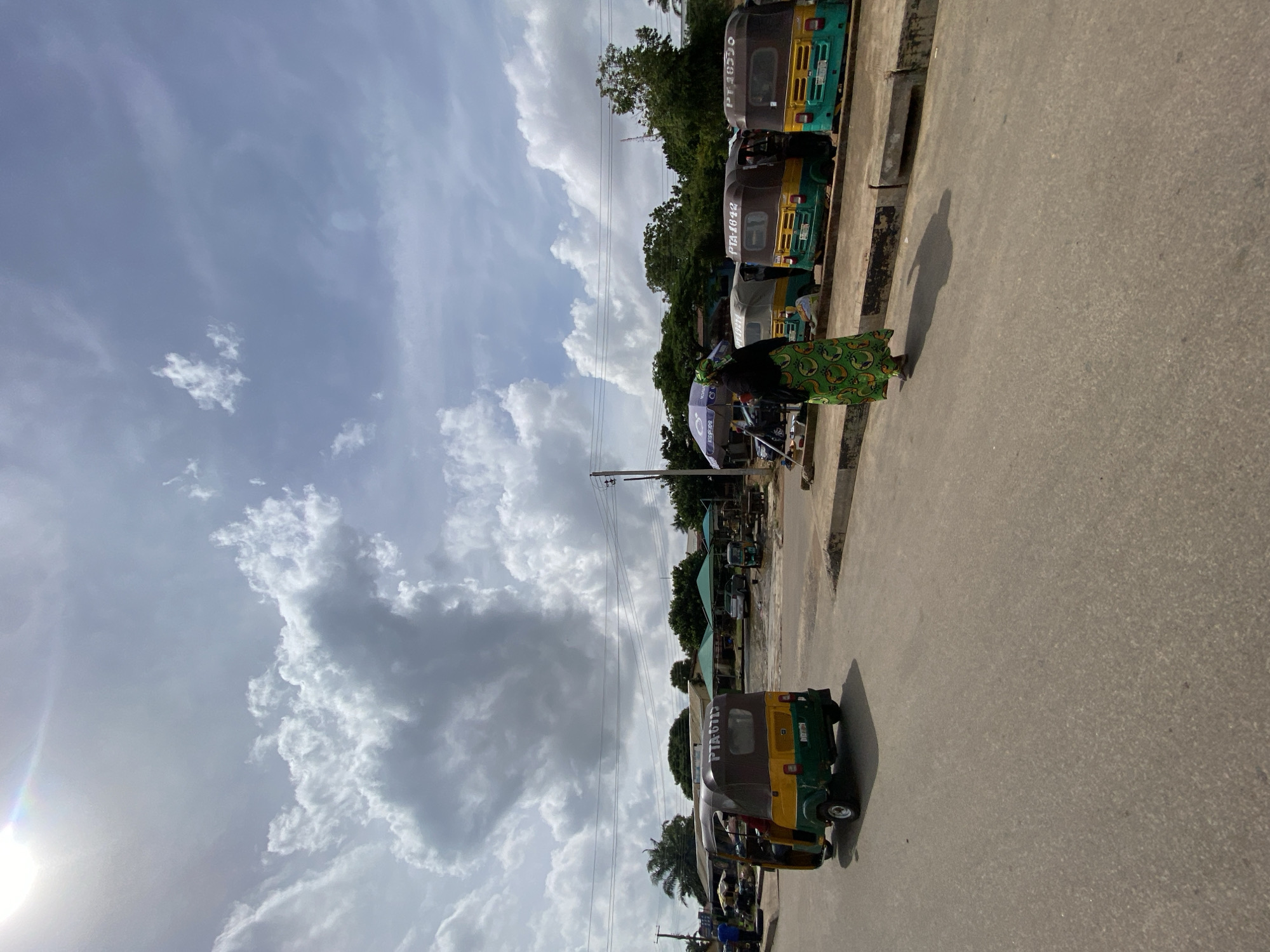 A wide shot captures an outdoor scene in Jos, Nigeria, under a partly cloudy sky. In the foreground, a paved road dominates the lower half of the frame. To the left, a yellow and green auto-rickshaw, a common mode of transport in Nigeria, is in motion, its rear facing the camera. Further ahead, parked along a curb, are several auto-rickshaws of similar design, some with visible license plates. A woman in a vibrant green patterned dress and headscarf walks along the curb, facing away from the camera. Behind the parked auto-rickshaws, several green and white canopies offer shade, suggesting a market or waiting area. Trees form a green backdrop to the scene, and utility poles with overhead wires are visible. The sunlight creates shadows on the road, indicating it is daytime. The overall impression is of a bustling, everyday street scene in a Nigerian city.