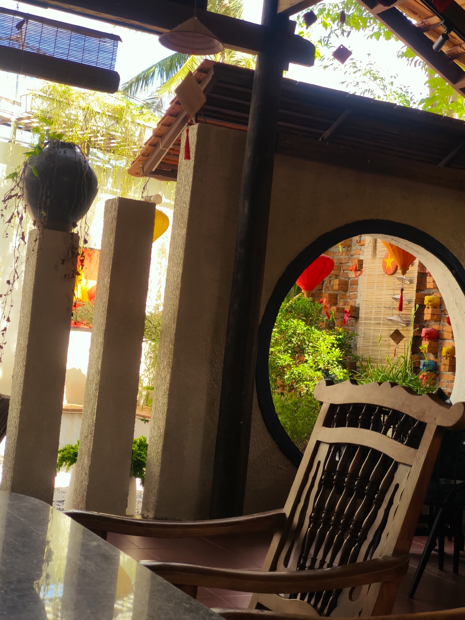 The image depicts an outdoor seating area at a cafe in Cam Ranh, Vietnam. In the foreground, a polished dark grey granite table occupies the bottom left corner. To the right, a vintage wooden rocking chair with a slatted backrest is positioned prominently. Beyond the chair, a circular opening in a textured beige wall reveals a glimpse of the cafe's exterior. This exterior features a brick wall adorned with several decorative elements including red and orange paper lanterns, small hanging ornaments, and woven bamboo blinds. Lush green foliage is visible through the circular opening, suggesting a garden or outdoor space. The background of the cafe area is characterized by a wooden roof structure with exposed beams and corrugated metal roofing. Hanging from the roof are a traditional Vietnamese conical hat, along with other decorative items. Several tall, narrow beige pillars are arranged vertically in the mid-ground, adding to the architectural style. On the left side of the pillars, a large dark ceramic pot with hanging vines is visible. The overall scene is bathed in natural light, indicating it was taken during the daytime.