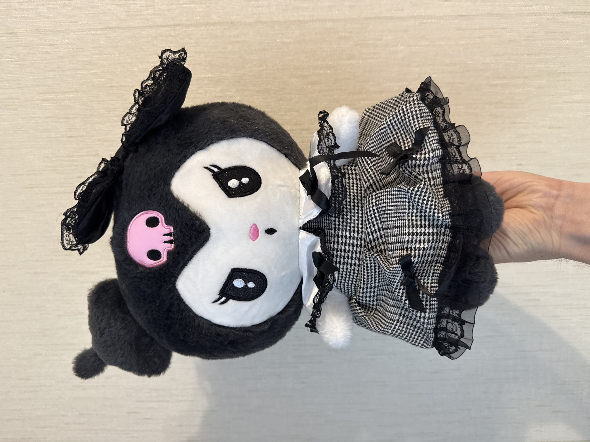 The image shows a plush toy, likely a character from a cartoon or anime, being held by a hand. The toy is predominantly black and white with a pink skull emblem on its cheek. It has large, expressive eyes and floppy ears adorned with black lace. The toy is wearing a black and white checkered dress with lace trim and black bows. The hand holding the toy appears to be Caucasian. The background is a light-colored, textured surface, possibly a wall or a piece of furniture. The lighting is bright, suggesting an indoor setting with ample light. The image is taken from a slightly angled perspective, focusing on the toy and the hand. There is no visible text in the image. The overall impression is that of a close-up shot of a decorative item being displayed or handled.