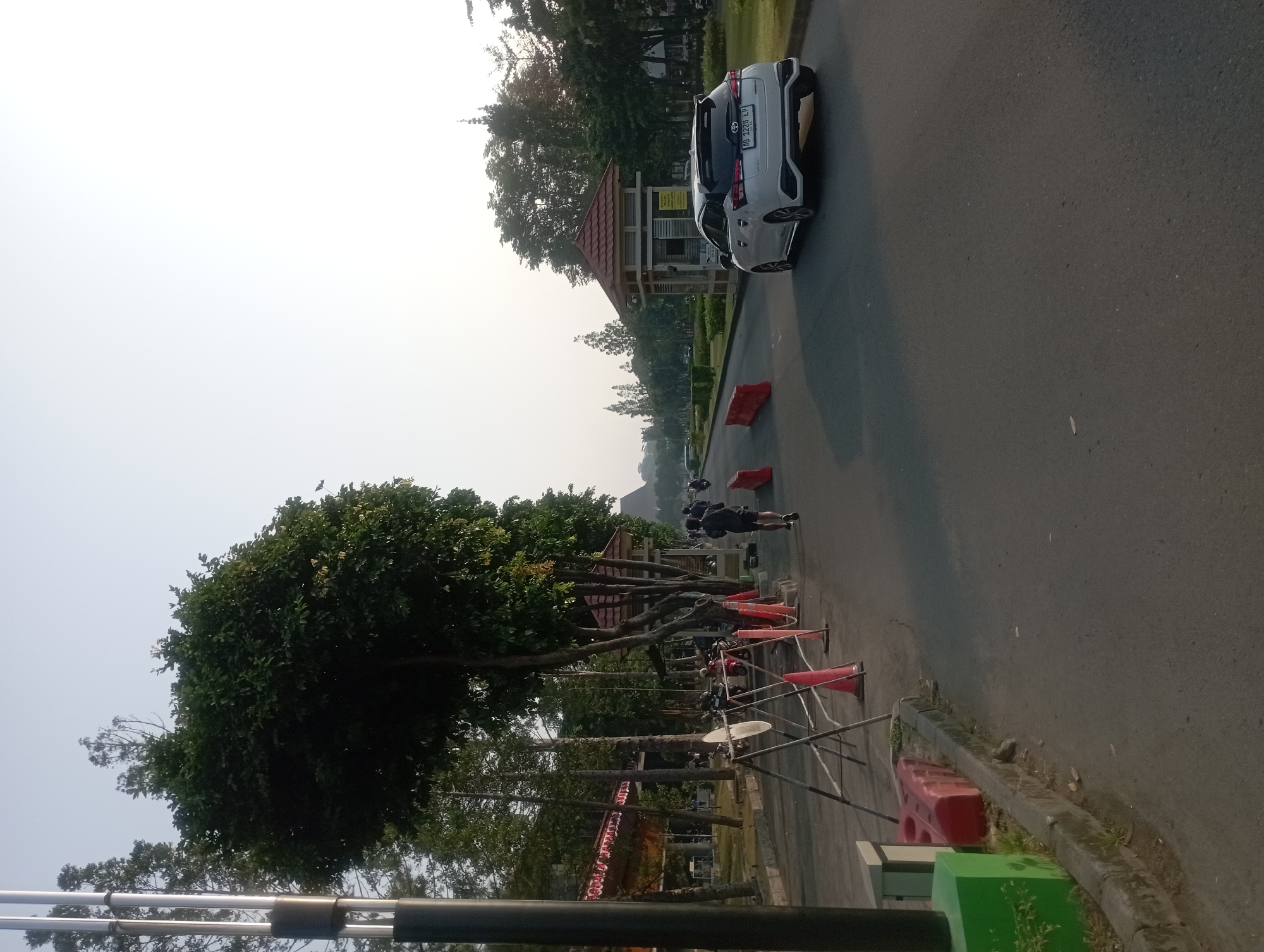 The image depicts a road in Depok, Indonesia, with a slight incline and lined with trees. The sky is clear and bright, indicating daytime, likely morning or late afternoon due to the quality of light. On the left side of the road, a large, lush tree dominates the foreground, with a tall metal pole beside it. Further down the road, a row of smaller trees and red-and-white striped awnings are visible, suggesting a building or facility. Barriers, including metal stands and orange traffic cones, are set up along the road's edge.
In the middle ground, two individuals are jogging on the right side of the road, wearing athletic attire. A white hatchback car is driving away from the viewer on the right side of the road, towards a small, elevated structure that appears to be a security post or gatehouse. This structure has a red tiled roof and a beige facade. In the background, more trees and buildings are visible, fading into the hazy distance. The overall scene suggests a public park, university campus, or a similar outdoor recreational area. The presence of joggers and a car suggests it's a functional space accessible to both pedestrians and vehicles. The white car has a license plate that reads "AB 1228 EP".