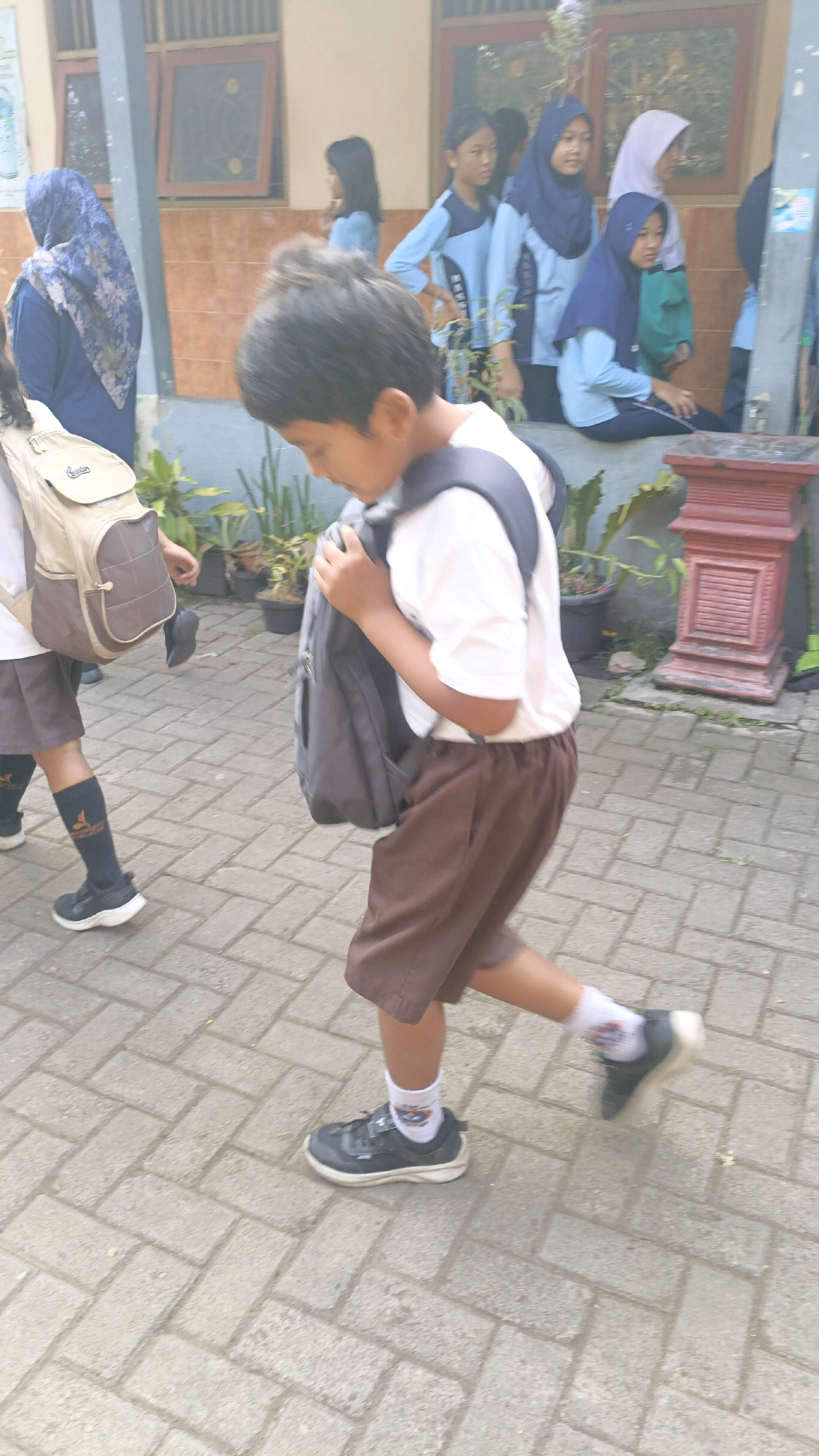 In Salatiga, Indonesia, on a sun-drenched day, a young boy with dark hair walks along a paved walkway in front of a building. He wears a white t-shirt and brown shorts, with black shoes and white socks. A dark backpack is slung over his shoulders. Other children, some wearing blue uniforms and headscarves, are visible in the background near the building. Plants are potted near the walkway, and a decorative red planter is present on the right. The scene appears to be a school environment, with students possibly heading to or from classes.
