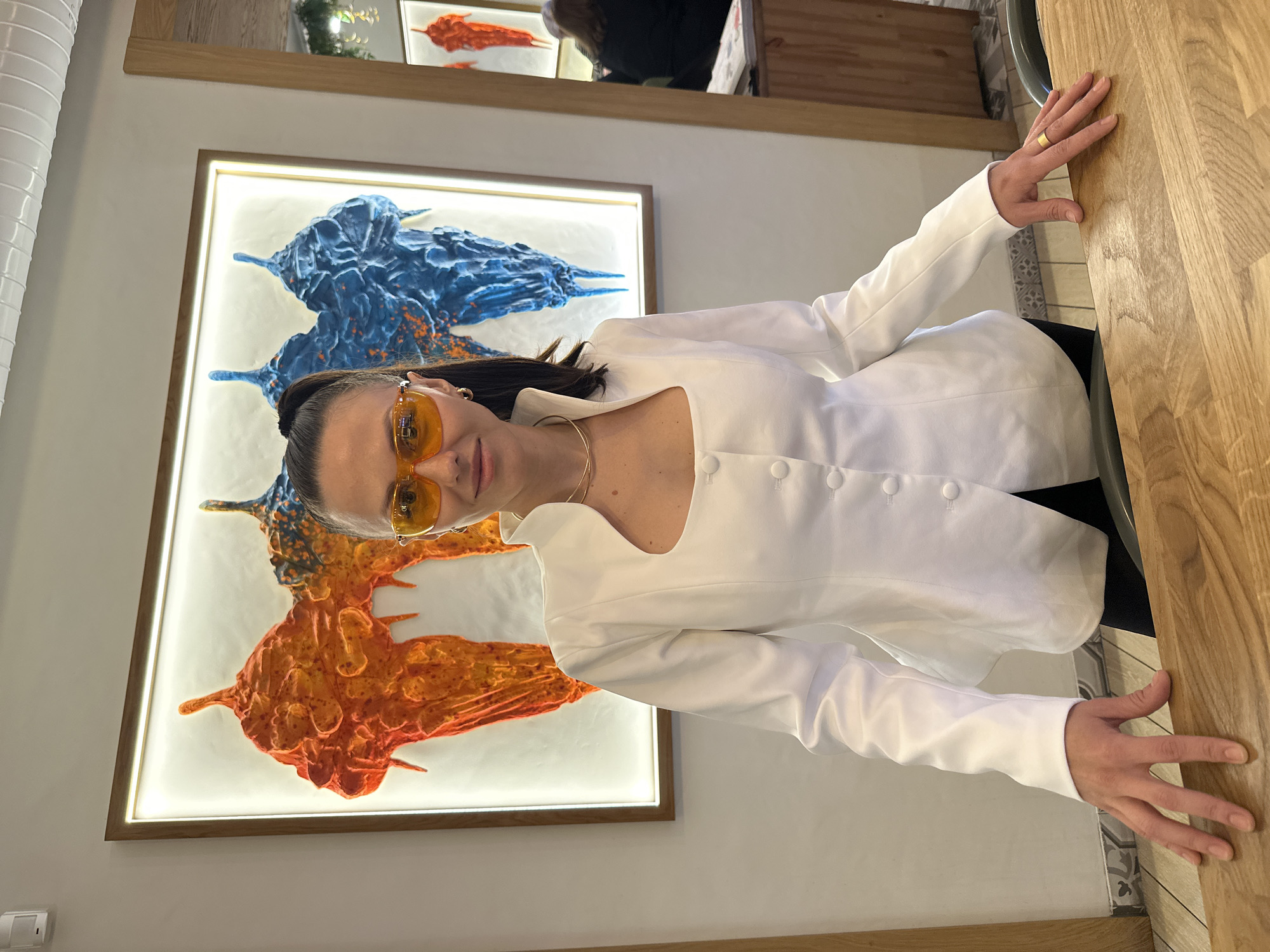 The image depicts a woman in a white, long-sleeved jacket and yellow-tinted sunglasses. She is standing near a wooden surface, possibly a table or countertop. Behind her is a framed piece of art with an illuminated background. The artwork displays two abstract, amorphous shapes, one predominantly blue and the other orange. The setting appears to be indoors, potentially a restaurant or gallery. Based on the presence of similar interior design elements, the location may be in Saint Petersburg, Russia, but the exact establishment is not identifiable.