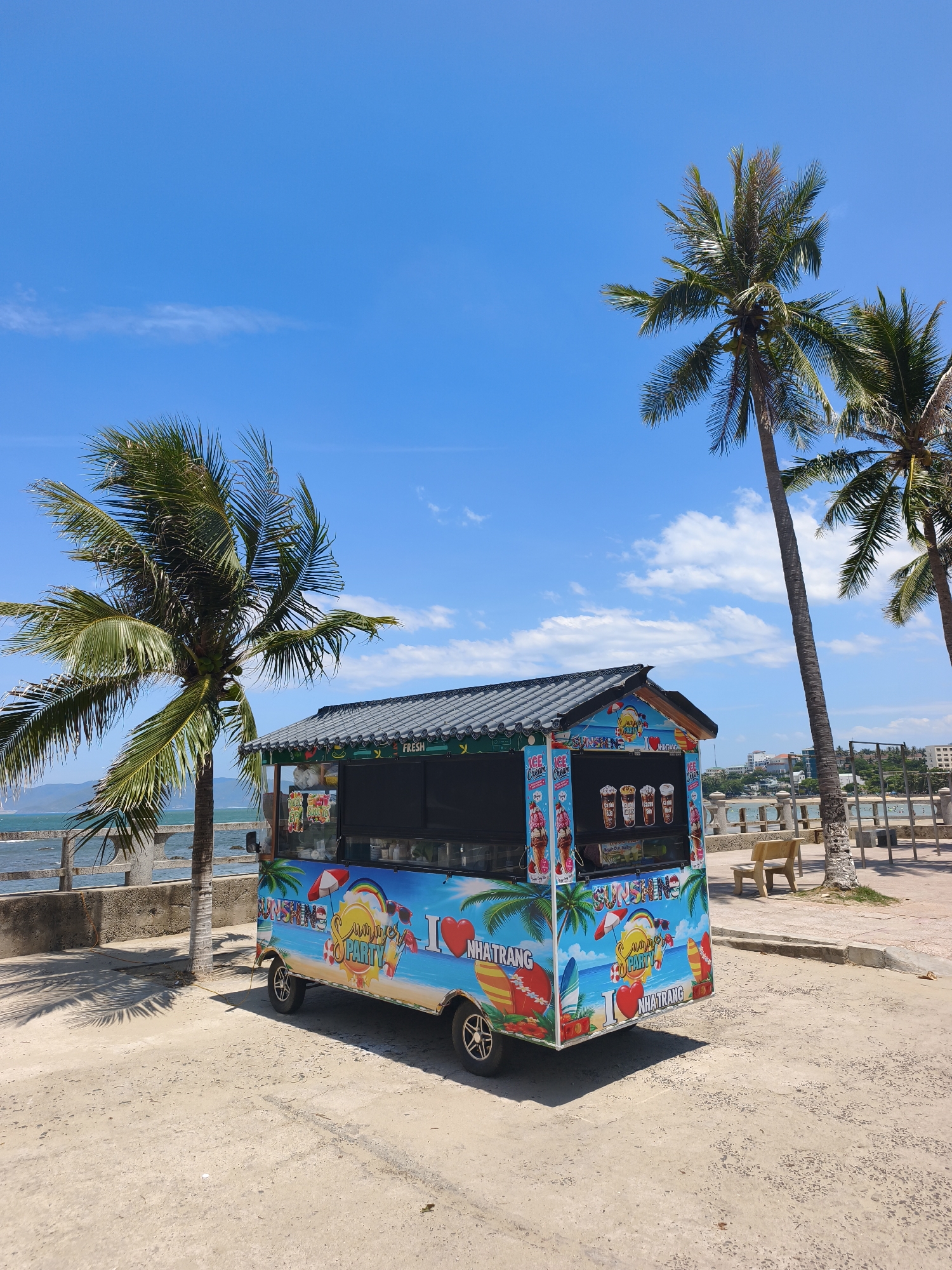 The image depicts a brightly colored mobile food cart parked on a sandy and paved area, likely a promenade or beachside walkway, under a clear blue sky with scattered clouds. The cart is adorned with tropical-themed graphics, including palm trees, surfboards, and the words "SUNSHINE SUMMER PARTY" and "I LOVE NHATRANG." It appears to be selling ice cream and beverages, as indicated by the depictions of ice cream cones and drinks displayed on the cart.
In the background, two tall palm trees frame the scene. Beyond the cart and trees, a concrete barrier separates the walkway from the sea, with the ocean visible stretching to the horizon. A section of a city or town, characterized by buildings and infrastructure, is also visible in the distance, suggesting an urban coastal setting. A light-colored wooden bench is positioned on the paved area near one of the palm trees. The overall environment evokes a sunny, vacation-like atmosphere typical of a seaside resort town. There are no people visible in the image. The image is taken from a slightly low angle, looking up at the cart and the sky.