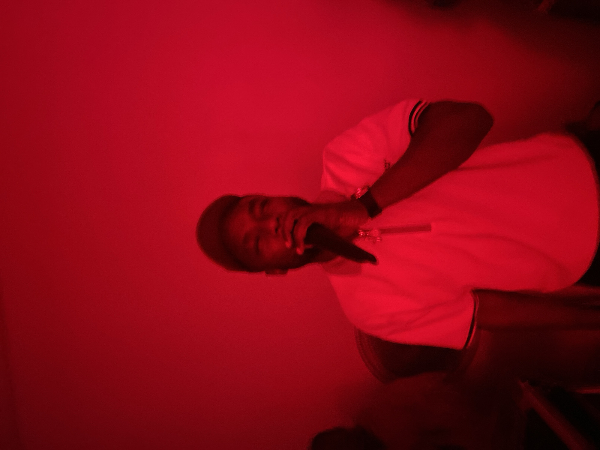 The image shows a person holding a microphone in front of their face, under red lighting. The person is wearing a light-colored polo shirt with dark trim on the collar and sleeves, and a baseball cap. They have dark skin and are looking towards the microphone. In the background, a blurred figure can be seen to the left, also under red lighting. The environment appears to be indoors, with a plain red wall behind the subjects.