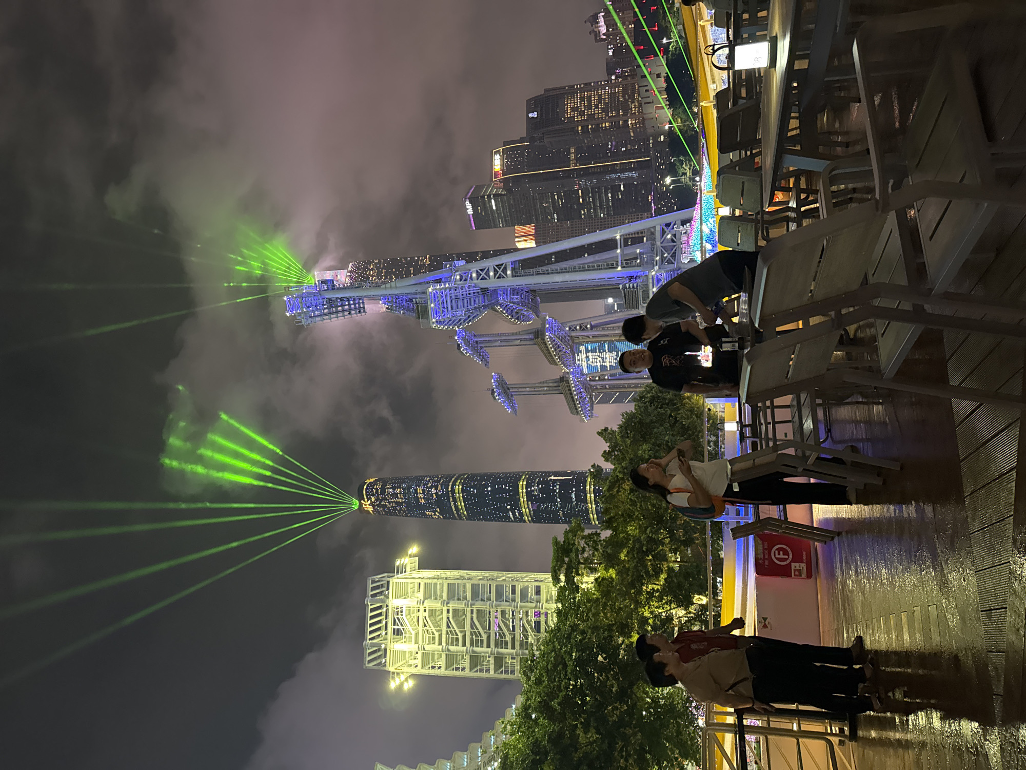 At night in Tianhe District, China, the Canton Tower is brightly lit with green lasers. A Ferris wheel is illuminated in blue. People stand and sit at tables on a balcony overlooking the scene. Other buildings are visible in the background. The weather appears foggy.