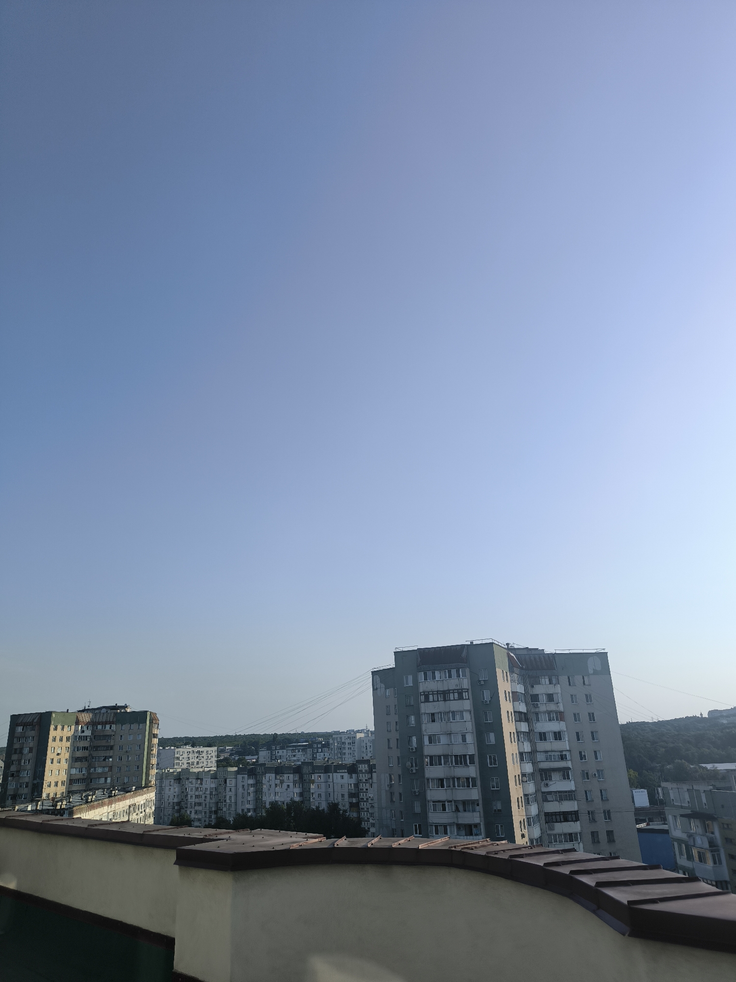 The image shows a view of an urban landscape in Chișinău, Moldova. In the foreground, there is a cream-colored wall with a brown, tiled edge, possibly a rooftop or balcony railing. Beyond this, a cluster of multi-story apartment buildings dominates the middle ground and background. The buildings are of varying heights and architectural styles, typical of Soviet-era housing blocks, with light-colored facades and numerous windows. Some of the buildings have visible air conditioning units. In the distance, a verdant hillside covered in trees can be seen, suggesting the city's location within a hilly terrain. The sky is a clear, pale blue, indicating it is likely daytime, with the sun casting gentle light on the scene. There are no people or vehicles immediately visible in the frame. The overall impression is one of a residential area within a city.