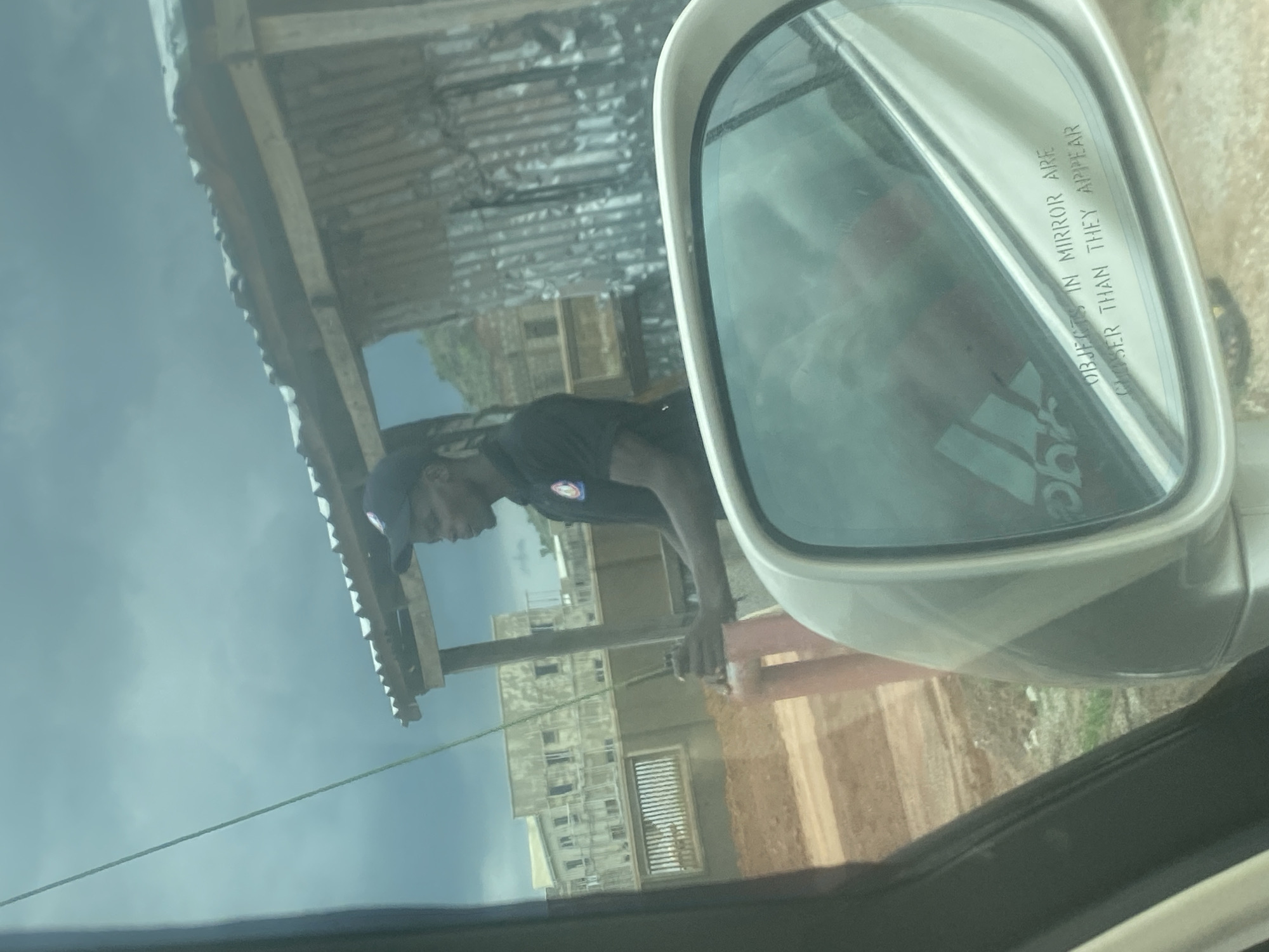 The image, taken from inside a vehicle, captures an outdoor scene in Gosa, Nigeria, rotated 90 degrees counter-clockwise. The primary subject is a dark-skinned male, wearing a dark cap and a dark t-shirt with a circular badge or logo on the left chest. He is positioned in the midground, leaning forward with his hands near a red vertical pole or pipe, seemingly engaged in an activity like working or observing.
In the foreground, the car's passenger-side mirror is prominent on the right, reflecting a blurred sky and a logo resembling the Adidas three stripes. The mirror's common safety warning "OBJECTS IN MIRROR ARE CLOSER THAN THEY APPEAR" is clearly visible in uppercase text.
Through the car window and beyond the person, a simple structure with a wooden frame and a corrugated metal roof is visible. Further in the background, several multi-story buildings, appearing to be made of concrete and possibly under construction, stand against a light blue or hazy sky. A dusty, reddish-brown dirt ground or path is seen below these structures. A thin rope or cable stretches horizontally across the lower-mid section of the frame. The scene takes place during daylight hours, under what appears to be an overcast or slightly hazy sky. The overall setting suggests a semi-urban or developing area.