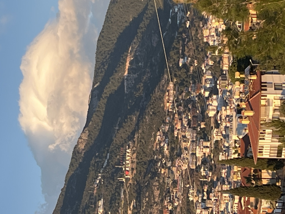 The image displays a panoramic view of the city of Alanya, Turkey, situated against a mountainous backdrop under a partly cloudy sky.
In the foreground, the upper sections of several residential or commercial buildings are visible, featuring tiled roofs predominantly in orange and terra cotta hues. Some rooftops are equipped with solar water heaters and chimneys. Tall, slender dark green trees frame the lower left and right edges of the frame.
The midground reveals a densely developed urban area consisting of numerous multi-story structures, primarily light-colored or white, extending across the undulating terrain at the base of a large mountain. A thin, dark cable or wire traverses horizontally across this urban expanse.
The background is dominated by a forested mountain range covered in dark green vegetation. Prominently displayed on a cleared section of the mountain slope is a large, white "I ❤️ ALANYA" sign, with the heart symbol rendered in red. Further up the mountain, exposed rock faces and less vegetated areas are discernible.
The sky in the upper portion of the image is blue, featuring a large, bright white cloud formation, likely a cumulus cloud, illuminated by sunlight from the side, suggesting late afternoon or early evening. Shadows are cast across parts of the mountain and urban areas, indicating the sun's angle.