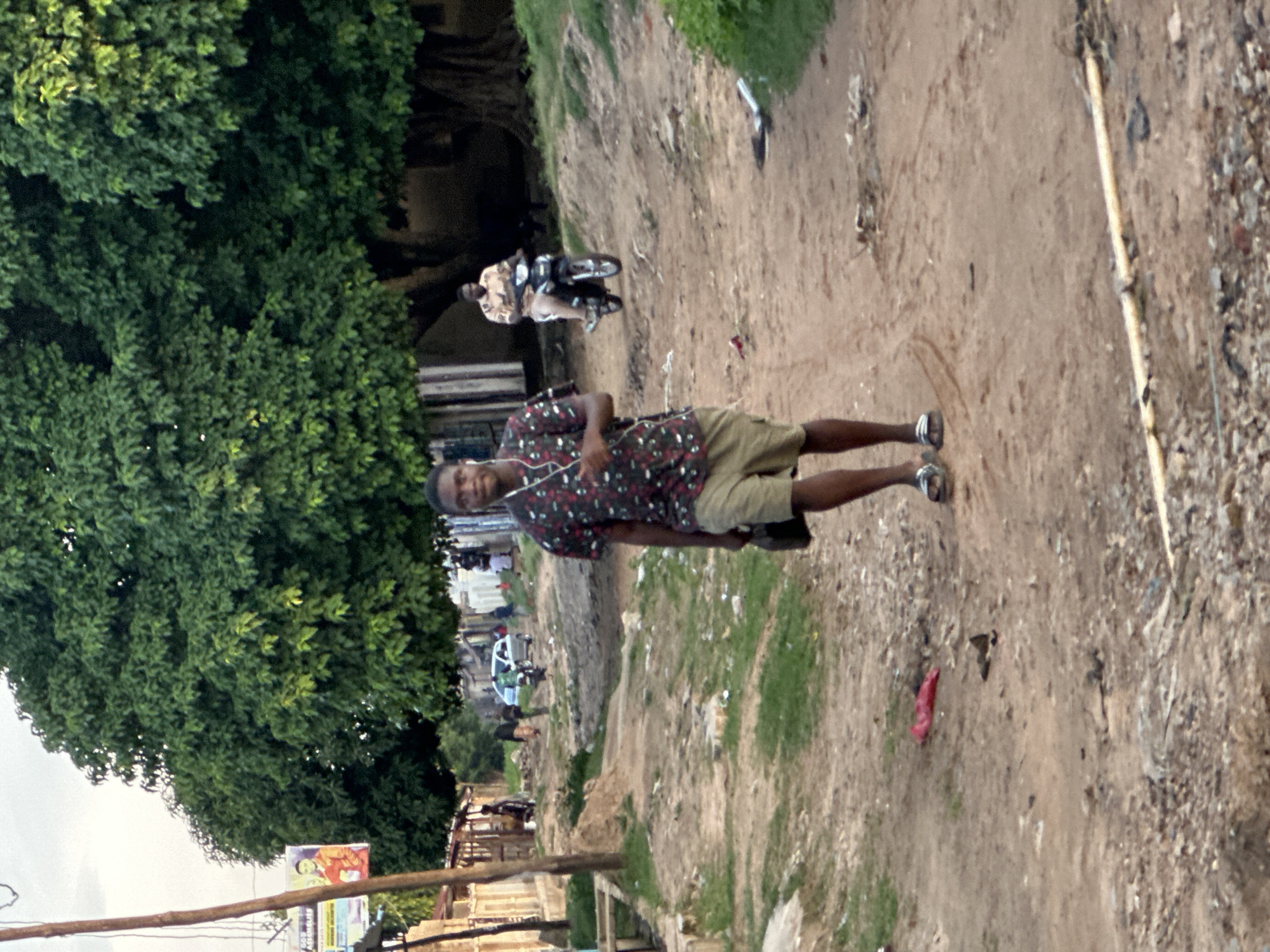 A young Black man wearing a patterned shirt and khaki shorts walks towards the camera on a dusty, unpaved road. He is carrying a dark bag in his right hand and has white earbuds in his ears. The road is lined with sparse vegetation and some scattered debris. In the background, a man rides a motorcycle and a white car is parked on the side of the road. There are buildings with corrugated metal roofs and trees with dense green foliage on either side of the road. A poster with an image of a person in an orange shirt is visible on a wooden pole to the left. The scene appears to be in a residential or semi-rural area of Narayi, Nigeria, with evidence of an informal, unpaved infrastructure and local life.