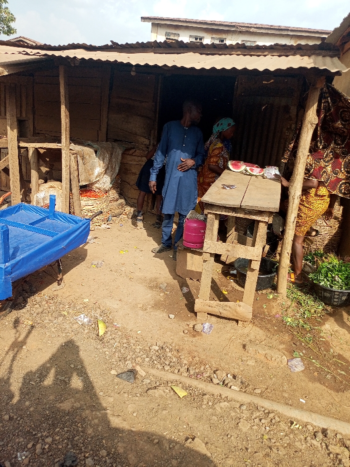 The scene depicts an outdoor market stall in Aramoko, Nigeria, under natural daylight. A rough wooden structure with a corrugated metal roof forms the stall, exhibiting walls constructed from unpainted planks and possibly corrugated sheets.
Centered, an adult male wearing a long-sleeved blue tunic stands, his left hand resting on his stomach. To his right, an adult female in a patterned dress and a light blue headscarf stands behind a rudimentary wooden counter, which displays two sliced watermelons. A pink gas cylinder is positioned on the ground adjacent to the counter. The lower portion of another individual, wearing a yellow patterned dress, is visible at the far right, standing near a black basin filled with green leafy vegetables. Inside the dim interior of the stall, large sacks of unspecified goods are discernible, along with a partially visible person wearing dark clothing on the left.
In the foreground, a blue metal pushcart or wheelbarrow with a fabric-lined bed rests on the dry, uneven dirt ground. The ground is littered with various debris, including plastic wrappers, paper, and organic waste, with a light green fruit rind visible near the center. Distinct shadows, including that of the photographer, are cast on the ground. A multi-story building with a light-colored facade is visible in the distant background, beneath a hazy sky.