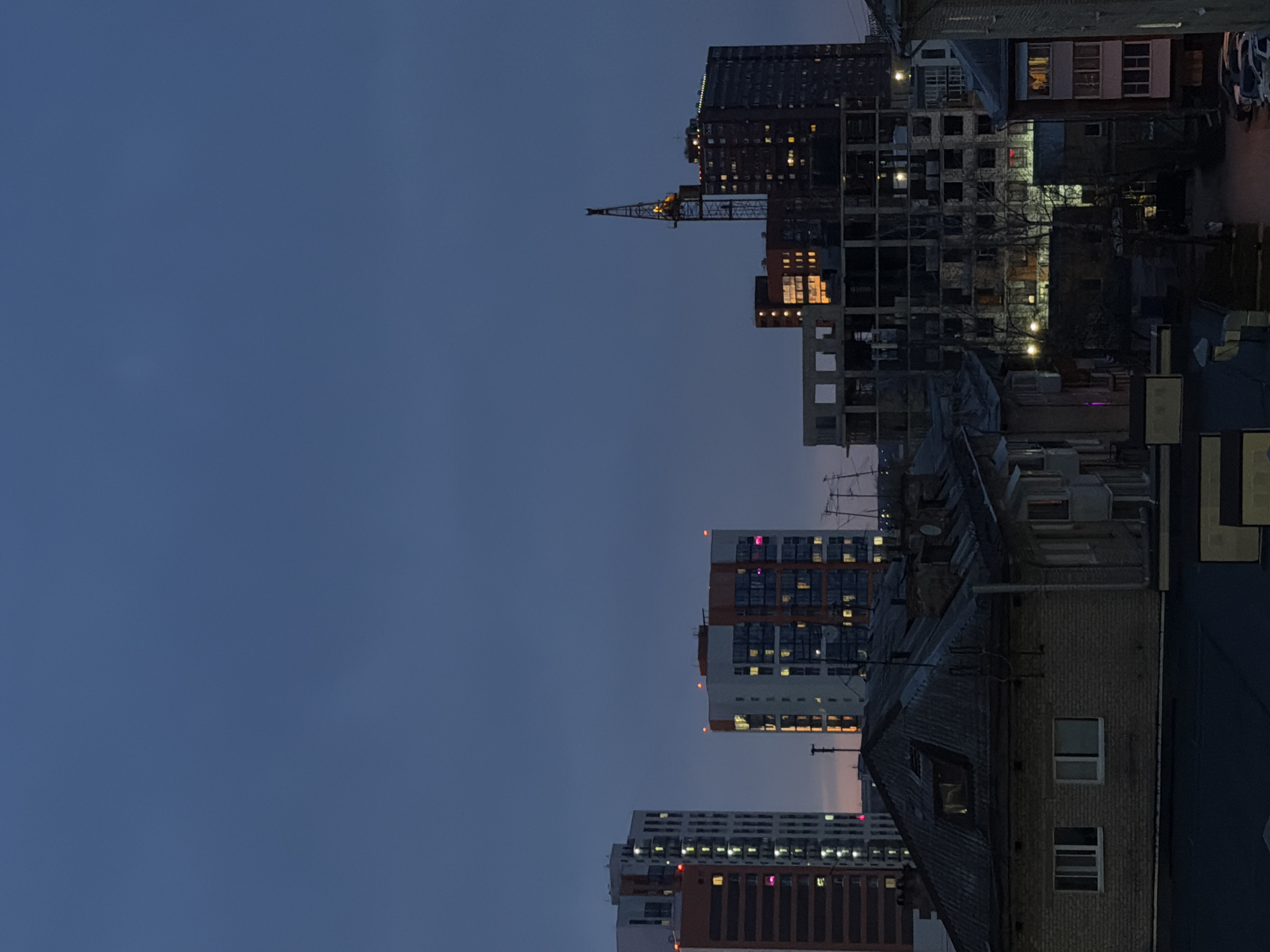 The image displays an urban cityscape at dusk or early night, viewed across various buildings under a dark blue, clear sky. The sky occupies the upper left and central portions of the frame, showing a deep indigo hue.
On the right side of the image, numerous buildings of differing heights and architectural styles are visible. A tall, brightly illuminated construction crane stands prominently in the mid-ground, suggesting ongoing development. Many windows across the buildings glow with interior lights, primarily warm yellow, with some exhibiting pink or magenta hues. One multi-story building in the mid-ground features a facade with white and orange-brown panels. Further back, taller and darker structures are present, some appearing as frameworks of buildings under construction.
In the foreground, the corrugated roof and brick walls of an older building are visible, also with several illuminated windows. Bare tree branches are discernible between the structures, suggesting a season without foliage. In the distant background, elements of a street or parking area, along with a parked vehicle, can be faintly seen. There are no people visible in the scene. The overall atmosphere is quiet and observant of city life transitioning into night. This urban vista is situated in Perm, Russia.