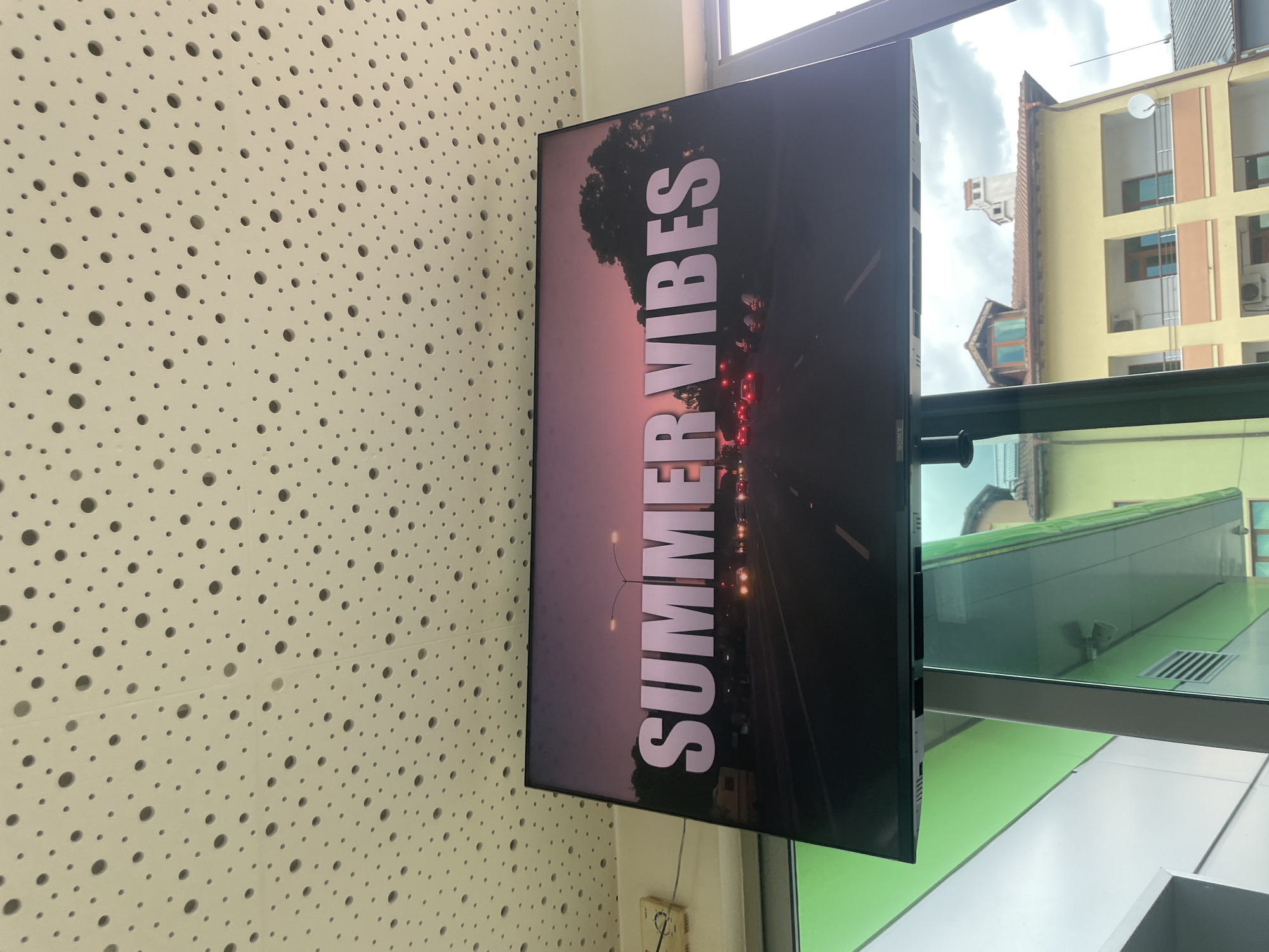 The image shows a television screen displaying the text "SUMMER VIBES" over a sunset road scene. The television is mounted on a ceiling with a perforated acoustic panel. To the left of the screen, there is a green wall and a device on the wall. In the background, visible through a window, are buildings with tiled roofs and balconies, indicating an urban or town setting. The sky is cloudy. The scene suggests an indoor commercial or public space in Ablanitsa, Bulgaria.