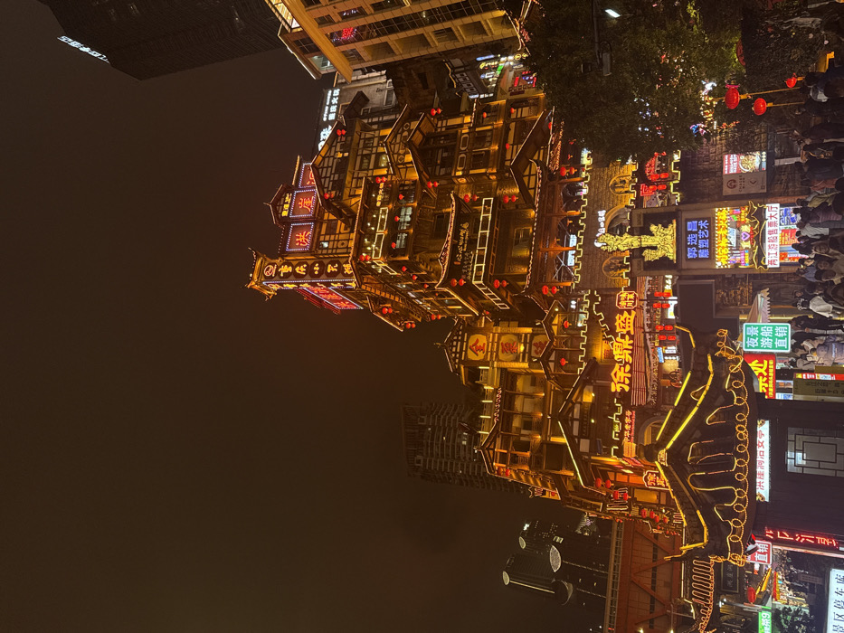 The image presents a nighttime scene in Yuzhong District, Chongqing, China, focusing on the densely illuminated Hongyadong complex. Multi-tiered traditional Chinese-style buildings, characterized by intricate roofs and balconies, are brightly lit with golden string lights outlining their structures and numerous red lanterns. Various neon signs displaying Chinese characters are visible on multiple levels of the buildings.
At the ground level and on lower platforms, a large crowd of individuals is observed congregating and traversing the area. A prominent golden statue or sculpture is situated above an arched entrance near the center-bottom of the primary architectural complex. Modern high-rise buildings appear in the dark background, partially obscured but visible above and to the sides of the traditional structures. The sky is dark and featureless, indicating nighttime conditions. Foliage from trees is visible on the right side of the lower foreground.