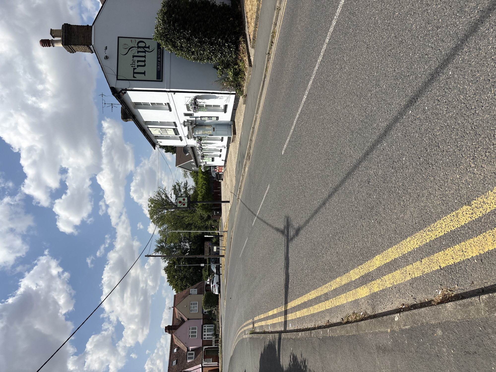 This is a ground-level, eye-level, outdoor, daytime, full-shot photograph of a street scene in Chelmsford, United Kingdom. The road in the foreground is asphalt, with double yellow lines painted on the left side of the carriageway. To the left of the road is a pavement with a curb. In the mid-ground, on the right side of the road, is a white two-story building with a pitched roof, identified by a sign as "the Tulip GRAY & SONS." This building has several windows, hanging flower baskets, and a light blue door. Further back, on the left side of the road, are several residential houses, painted in various colors including pink and beige, with pitched roofs and gardens. Trees and foliage are visible throughout the scene. The sky above is blue with scattered white cumulus clouds. There are no people visible in the photograph. The overall atmosphere is that of a sunny day in a suburban or semi-rural area.