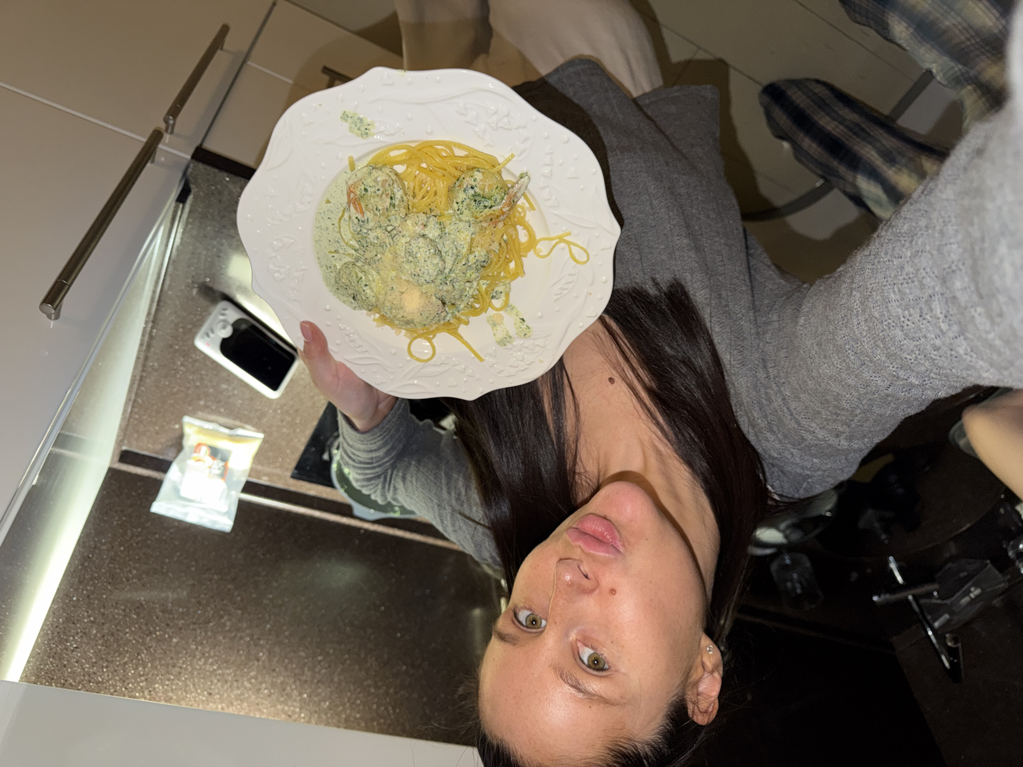An eye-level indoor shot captures a young woman holding a plate of food, looking directly at the camera. The woman, with long dark hair and green-brown eyes, wears a neutral to slightly pouty expression. She is dressed in a grey, ribbed long-sleeved top. Her left hand supports a decorative white plate with scalloped edges, filled with a portion of spaghetti topped with a vibrant green, creamy sauce and several pieces of what appear to be shrimp or meatballs, also coated in the sauce. Green herb flecks are visible on the food and plate.
The background indicates an indoor setting, likely a kitchen or dining area in Khimki, Russia, as per the provided context. A dark-colored countertop stretches behind the plate, holding a white device with a black screen and buttons, possibly a remote control or a compact camera. Further back on the countertop, a small transparent plastic bag containing indistinct items is visible. On the far left, two metallic rods are mounted horizontally, suggesting part of an appliance or cabinet hardware. Artificial lighting illuminates the scene, implying it could be evening or nighttime. A section of plaid fabric, possibly a blanket or clothing, is visible in the upper right quadrant of the image. The woman has a small stud earring in her visible ear. No clear text is discernible within the image. The overall activity portrays the woman presenting a homemade meal, possibly for a photograph.