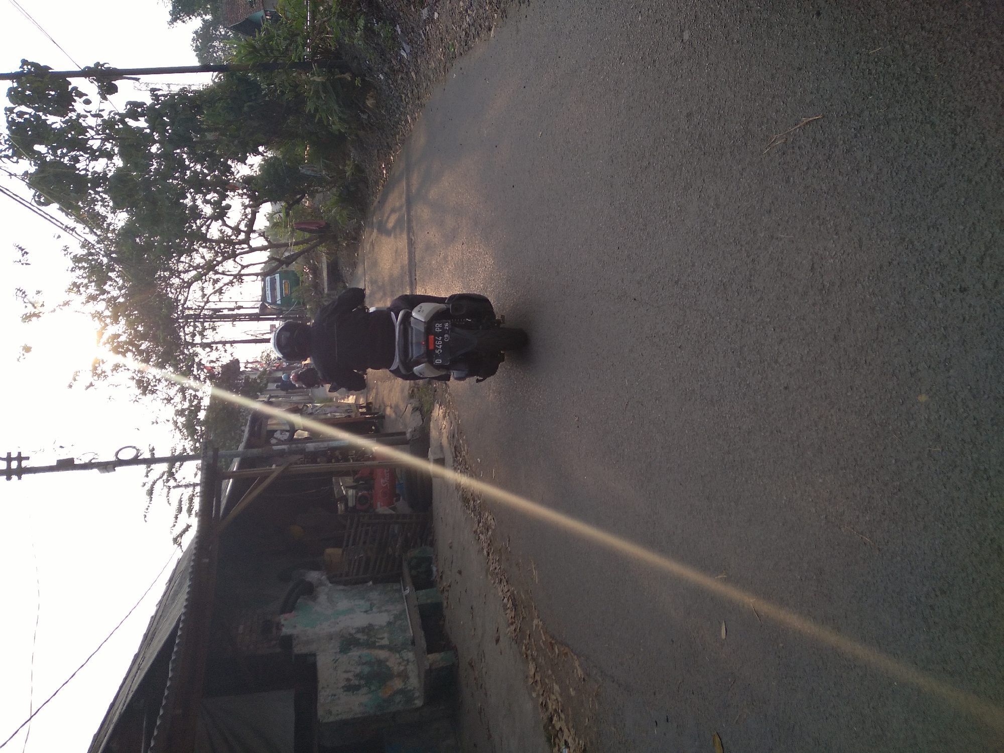 The image depicts a street scene in Cimahi, Indonesia, with a person riding a scooter from a rear perspective. The scooter has a license plate that reads "D 5464 PR 09 26". The rider is wearing a black jacket and a helmet. The road is asphalt, and there is vegetation and a simple building with a corrugated metal roof on the left side of the road. Power lines are visible overhead, and the sun is setting or rising, casting long shadows and creating a bright glare in the upper portion of the image. The environment appears to be a somewhat rural or suburban area.
