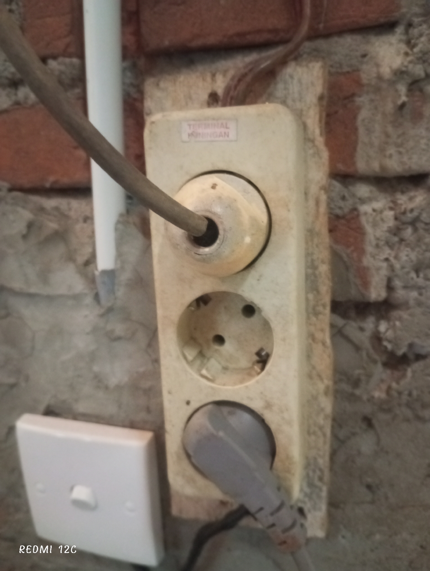 The image shows an old, dirty multi-outlet electrical socket mounted on a rough, brick wall. A cord is inserted into the top outlet, and a plug is inserted into the bottom outlet. A white light switch is located next to the socket. The location is in Lhokseumawe, Indonesia.