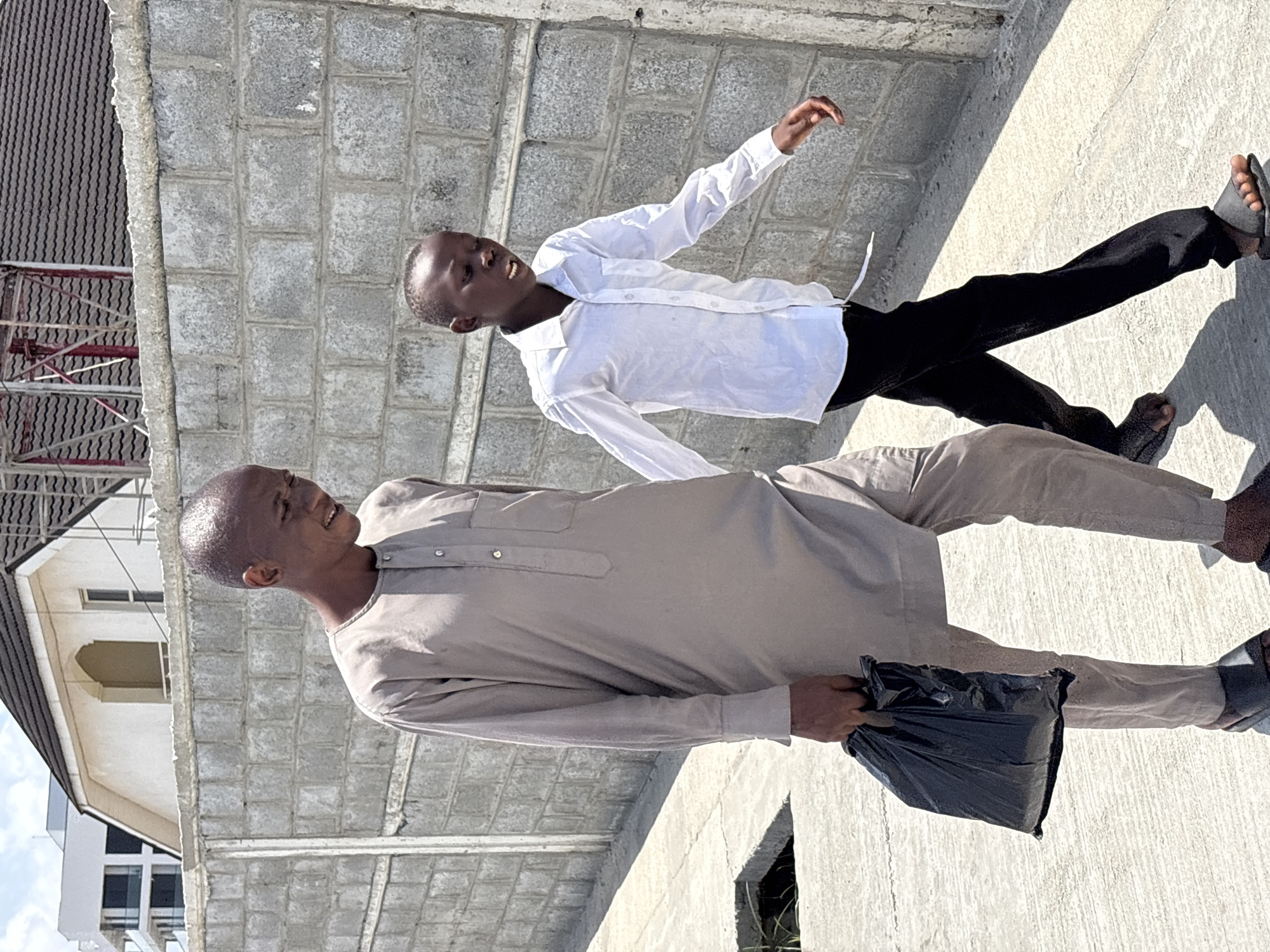 This image, taken in Alaguntan, Nigeria, features two smiling males outdoors on what appears to be a concrete rooftop or an unfinished upper floor of a building, under bright daylight.
The primary subject is an adult male with a shaved head and dark skin, dressed in a light brown or beige traditional tunic (possibly a kaftan or agbada style top) and matching trousers. He holds a black plastic bag in his left hand and is looking upwards and to his left with a wide smile.
Standing slightly behind and to his right is a younger male, a boy, also with dark skin and short hair. He is wearing a white long-sleeved button-up shirt and black trousers, with dark sandals. His right arm is raised and bent, and his mouth is slightly open in an animated expression, suggesting a lively moment or interaction. Both individuals appear happy and content.
The setting is characterized by rough, grey cinder block walls and a concrete ground, indicating an area under construction or a utilitarian rooftop. In the background to the left, part of a finished building with a dark tiled roof, white walls, and arched window details is visible, adding a local architectural element. Strong, distinct shadows are cast on the ground, typical of a sunny day around midday or early afternoon. No visible text is present in the image.