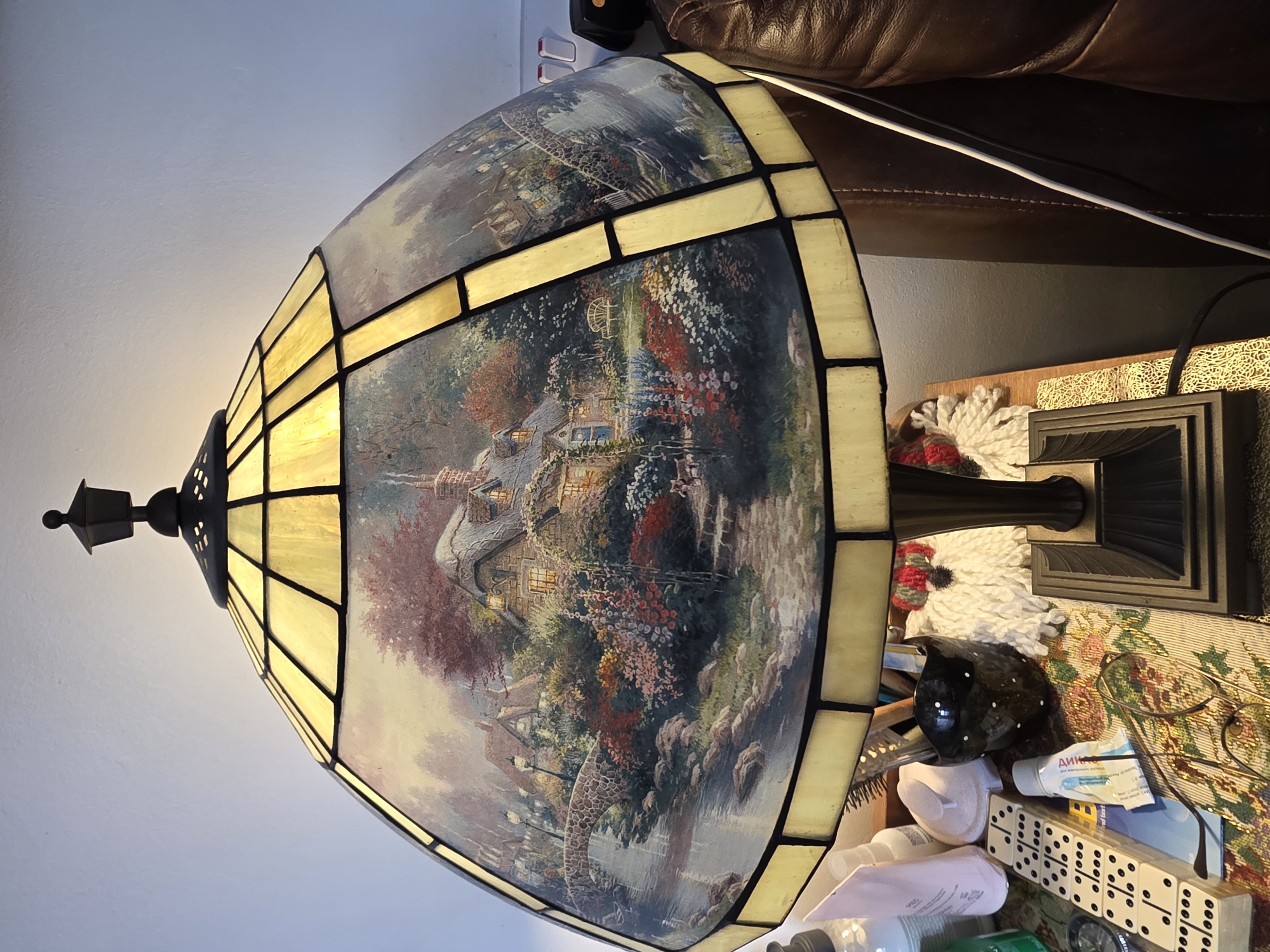 A large, illuminated Tiffany-style table lamp dominates the foreground, placed in an indoor setting in Cwmbrân, United Kingdom. The lampshade features numerous segmented panels of yellowish-orange stained glass interspersed with larger, painted glass panels. These painted sections depict idyllic, detailed landscapes, primarily featuring quaint cottages with glowing windows, lush gardens bursting with colorful flowers, arched bridges over water, and winding paths, reminiscent of a pastoral scene. The lamp's base, partially visible, is dark and appears to be made of metal or resin, with decorative fluting.
The lamp sits on a cluttered surface, likely a side table. Visible items on the table include a stack of dominoes, various bottles and tubes of toiletries or medicine (one partially showing "ДИКА" in Cyrillic, and another "and find tart"), a pair of reading glasses resting on a patterned fabric, a black glossy bird-shaped decorative object, and a white, textured, crocheted item.
In the background, a brown leather armchair or sofa is visible, with a white charging cable draped across it. A plain light-colored wall forms the upper left background. The lamp itself provides a warm, inviting glow, suggesting it is evening or that the room is dimly lit, contributing to a cozy and domestic atmosphere.