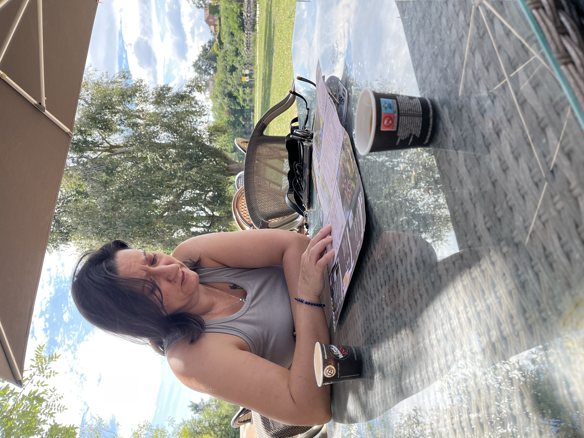 In Saint-Just-Saint-Rambert, France, a woman with dark hair is sitting at a glass-top table outdoors, looking at a menu. She is wearing a gray tank top and a blue beaded bracelet. Two small coffee cups are on the table, one in front of her and one to her right. The table is set on a patio with a large umbrella providing shade. In the background, trees, a body of water, and buildings are visible under a partly cloudy sky. A woven chair is behind the woman.