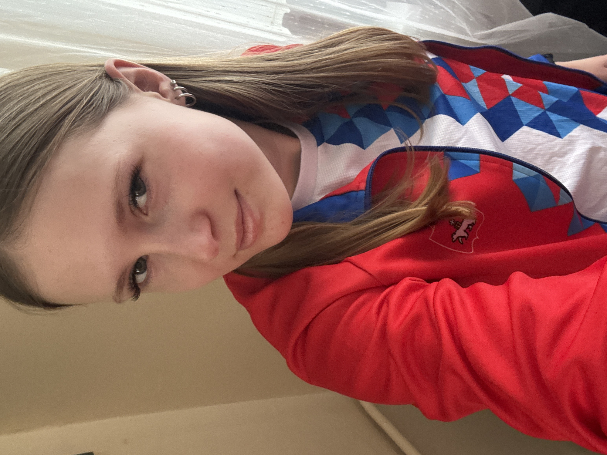 A portrait of a young woman with fair skin and light brown hair is captured in a close-up shot. She has light-colored eyes, a slight smile, and multiple piercings in her left ear. She is wearing a white top with a blue and white geometric pattern overlaid by a red jacket with a small, possibly embroidered, crest on the left side.
The background is indistinct, suggesting an indoor setting. The overall composition is simple, with the focus primarily on the woman and her attire. There is nothing in the image to suggest the location is Berezniki, Russia.