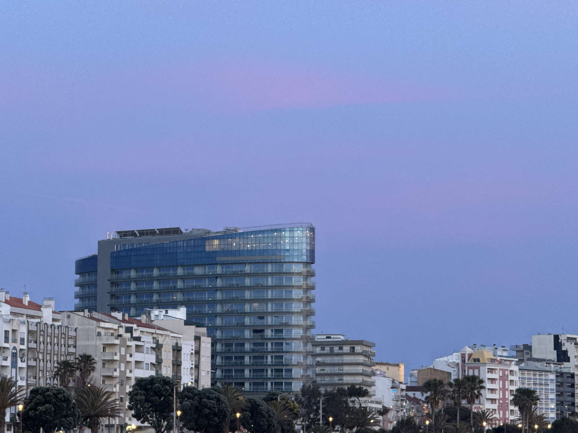 A view of buildings in Figueira da Foz, Portugal, at dusk. A modern, high-rise glass building with balconies dominates the center-left of the frame. Adjacent to it are several older, white-painted buildings with tiled roofs and balconies. Palm trees and streetlights are visible in the foreground, lining the street. The sky is a gradient of blue and pink, suggesting the time of day. No people are visible.