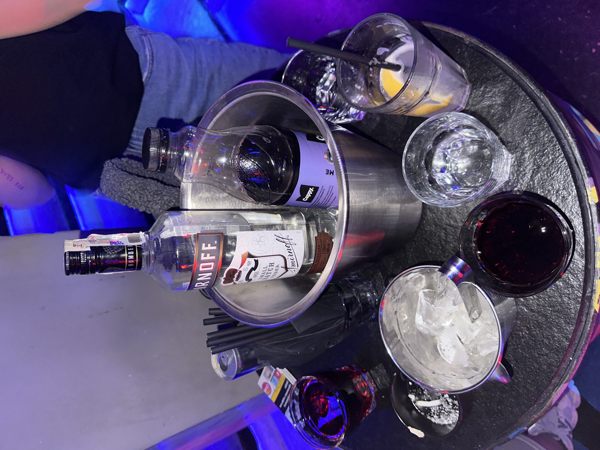 Here is a detailed description of the image:
**Scene:** The image is a top-down view of a small, black, round table in a bar or nightclub setting. There is a person wearing jeans and a black shirt visible in the upper left corner of the frame.
**Objects:**
* A metal ice bucket containing bottles of Smirnoff vodka and Cappy juice.
* Several black drinking straws are in the ice bucket.
* Drinks in various glasses, including a clear liquid, a dark liquid (possibly cola or red wine), and a mixed drink with lemon.
* A small dish or coaster with a white powdery substance (possibly salt or sugar).
**Environment:**
* The lighting is dim and has a noticeable blue hue, suggestive of club or bar lighting.
* The table is round and black, with a textured surface.
**Location:** Based on the brand of juice and vodka, it is possible that the scene could be in a bar or nightclub in the city of Varna, Bulgaria.
