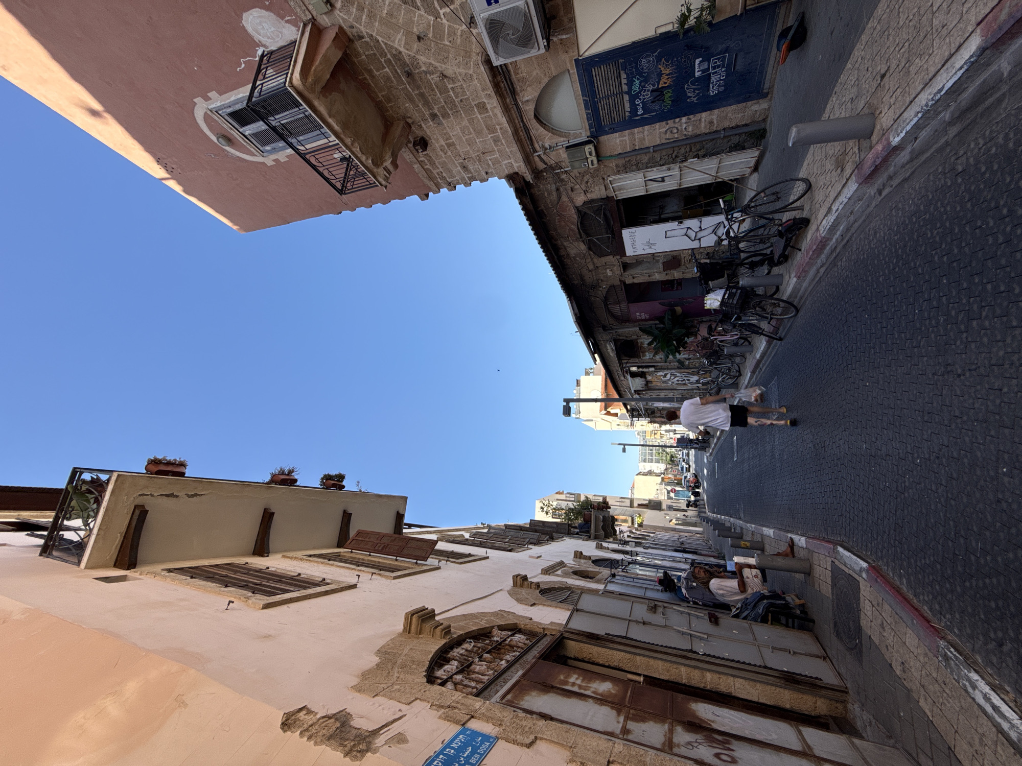 The image captures a sun-drenched, narrow street in Tel Aviv, Israel, likely in an older district given the architecture. The street is paved with dark cobblestones and flanked by buildings of varying architectural styles. On the left, a light-colored building with weathered plaster displays multiple levels, featuring arched doorways, barred windows, and balconies adorned with potted plants. A blue street sign with Hebrew lettering is affixed to its facade. On the right, a building constructed from rough-hewn stone rises, its upper portion painted a reddish-pink. This building also has a balcony and an air conditioning unit mounted on its facade. Further down the street, shops with open doors and metal shutters line the pavement, some with signage. A single person, a man wearing a white t-shirt and black shorts, walks away from the camera, carrying a plastic bag. Another person, appearing to be a man with long hair, sits on a low wall on the left side of the street. Several bicycles and a scooter are parked along the sidewalk. The sky is a clear, vibrant blue.