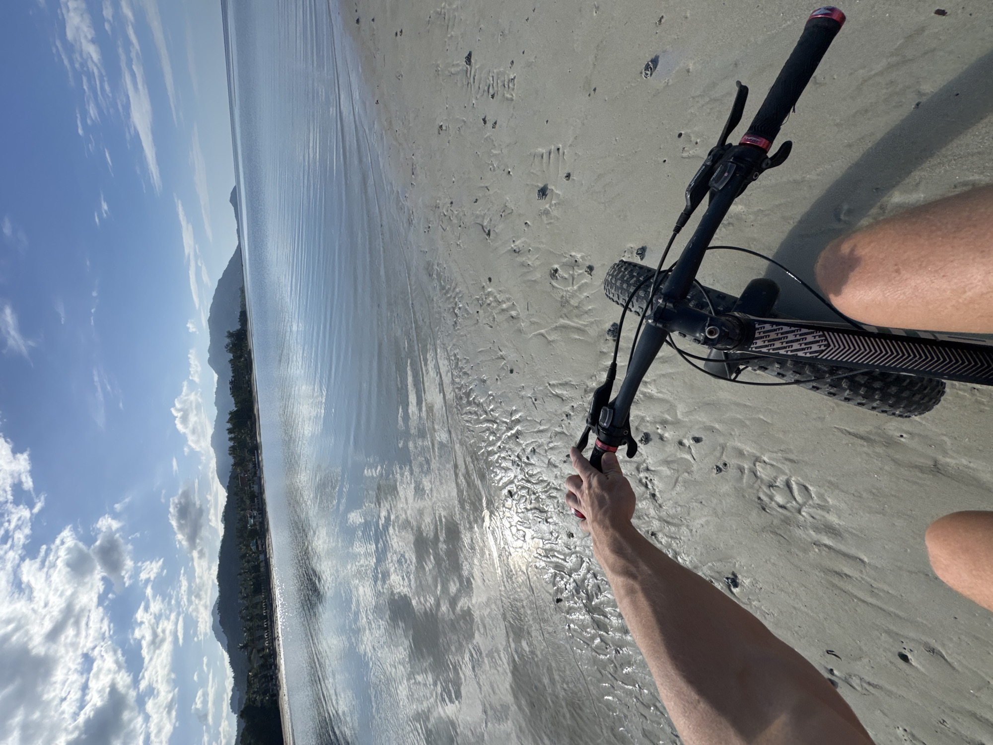 The image shows a first-person perspective of someone riding a fat-tire bicycle on a sandy beach. The rider's left arm and hand are visible, gripping the handlebars. The bicycle is black with distinctive patterned frame tubes. The beach is wet and sandy, with ripples and some scattered debris. The ocean stretches out to the horizon, reflecting the blue sky and white clouds. In the distance, a line of lush green hills forms the coastline, with buildings and palm trees visible along the shore. The scene appears to be set in a tropical location, likely ตำบลเกาะพะงัน, Thailand, given the natural landscape and the clear blue sky. The lighting suggests it is daytime, possibly morning or late afternoon, with the sun casting a gentle glare on the water and sand.