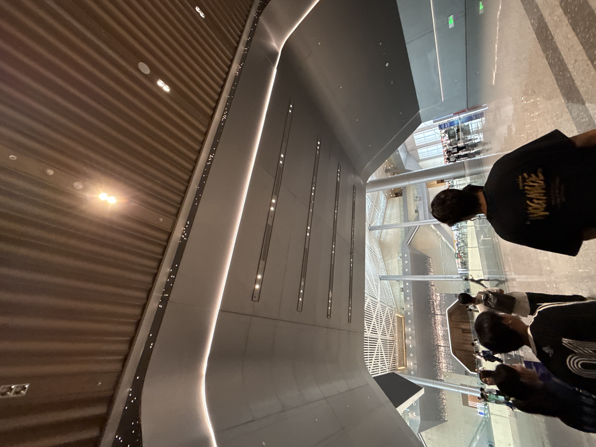 The image shows the interior of a modern building with a high ceiling. The ceiling is made of wood panels with integrated lighting. The walls are curved and made of a smooth, light-colored material, with embedded linear lights. In the foreground, a group of people are walking away from the camera. One person in the center is wearing a black t-shirt with yellow text that reads "YOG MADE". The floor is reflective, possibly polished concrete or marble, and shows reflections of the ceiling lights and the architecture. In the background, a large, open space with escalators and multiple levels is visible. The overall impression is of a spacious and contemporary public building, likely an airport terminal, train station, or convention center, located in the Chongqing High-tech Zone, China.