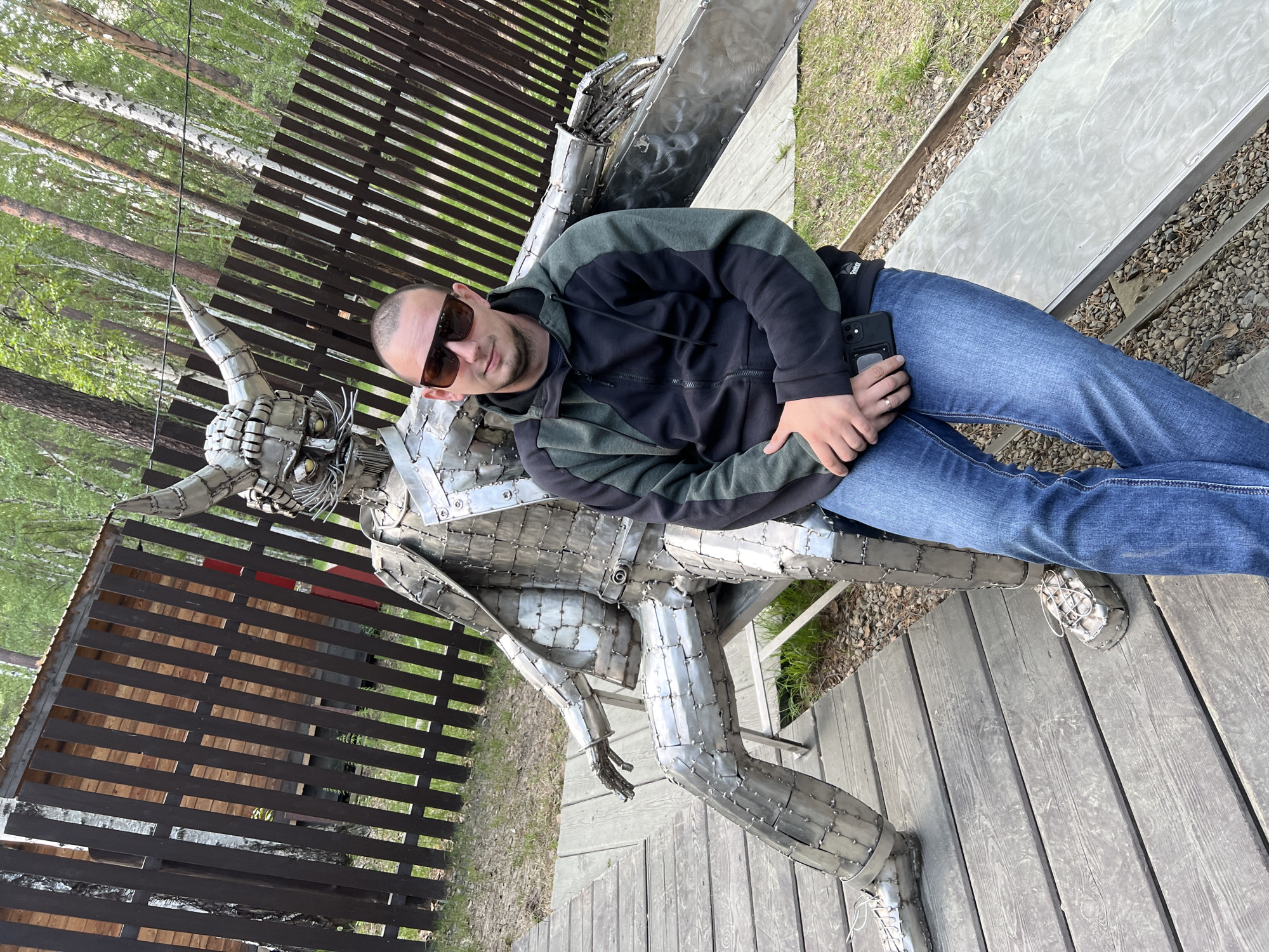 In Tal'tsy, Russia, a man in sunglasses and a black and green zip-up hoodie, blue jeans, and sneakers sits on a metal bench next to a large, detailed metal sculpture of a horned, armored figure. The sculpture is made of many small metal pieces riveted together, forming a seated warrior with a helmet, cape, and metal shoes. The man holds a black smartphone in his right hand. They are positioned on a wooden boardwalk in an outdoor setting with a wooden fence and trees visible in the background. The ground around the boardwalk is covered in small stones and grass.