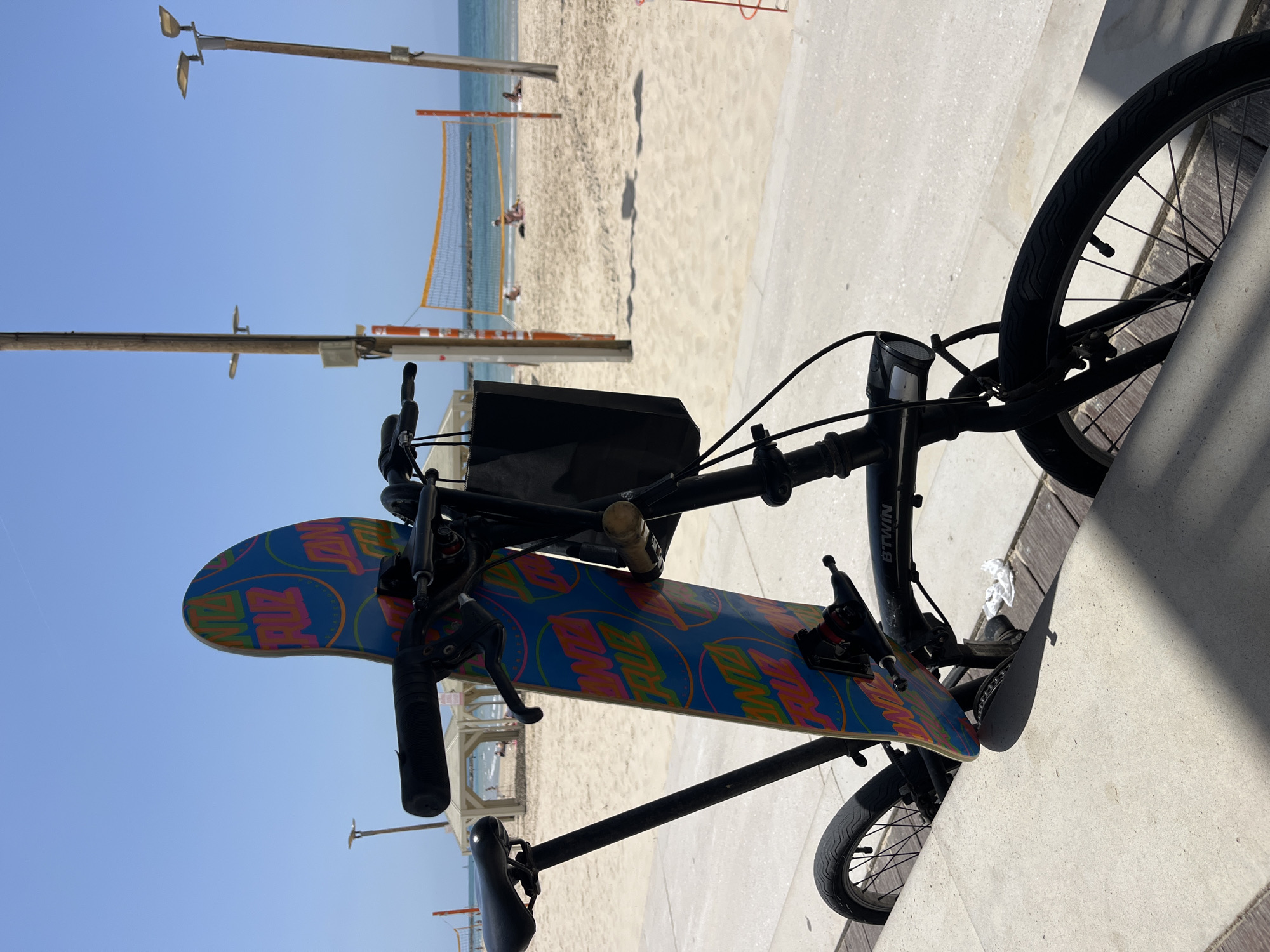 A bicycle with a skateboard attached to the handlebars is situated on a concrete promenade adjacent to a beach. The bicycle is black and has a "BTWIN" brand label visible on the frame. A black bag or container is attached above the handlebars. The skateboard is blue with colorful Santa Cruz logo patterns and features black trucks and wheels. The beach in the background has volleyball nets and people. Light posts are visible along the promenade. The location appears to be Tel Aviv, Israel, based on the beach and promenade architecture.