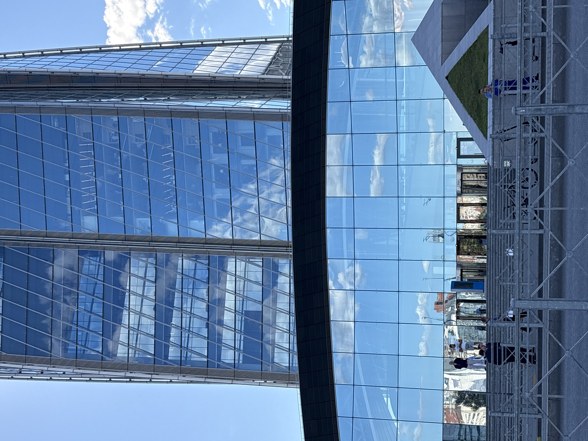 The image depicts a modern skyscraper with a glass facade, reflecting the sky and clouds. The building is made of numerous glass panes arranged in a grid pattern. Part of the building's structure, including window frames and horizontal supports, is visible. In the foreground, there is a metal scaffolding structure. On a small grassy area adjacent to the building, a person wearing a blue shirt and dark pants is standing. Reflected in the glass facade are other buildings and the sky. Several people are visible as reflections on the glass, some standing and some walking. The scene appears to be outdoors during the daytime, with a clear blue sky and scattered white clouds. The overall setting is urban, likely a business or financial district, given the architecture of the skyscraper. There is no visible text in the image. The orientation of the image is rotated 90 degrees clockwise.