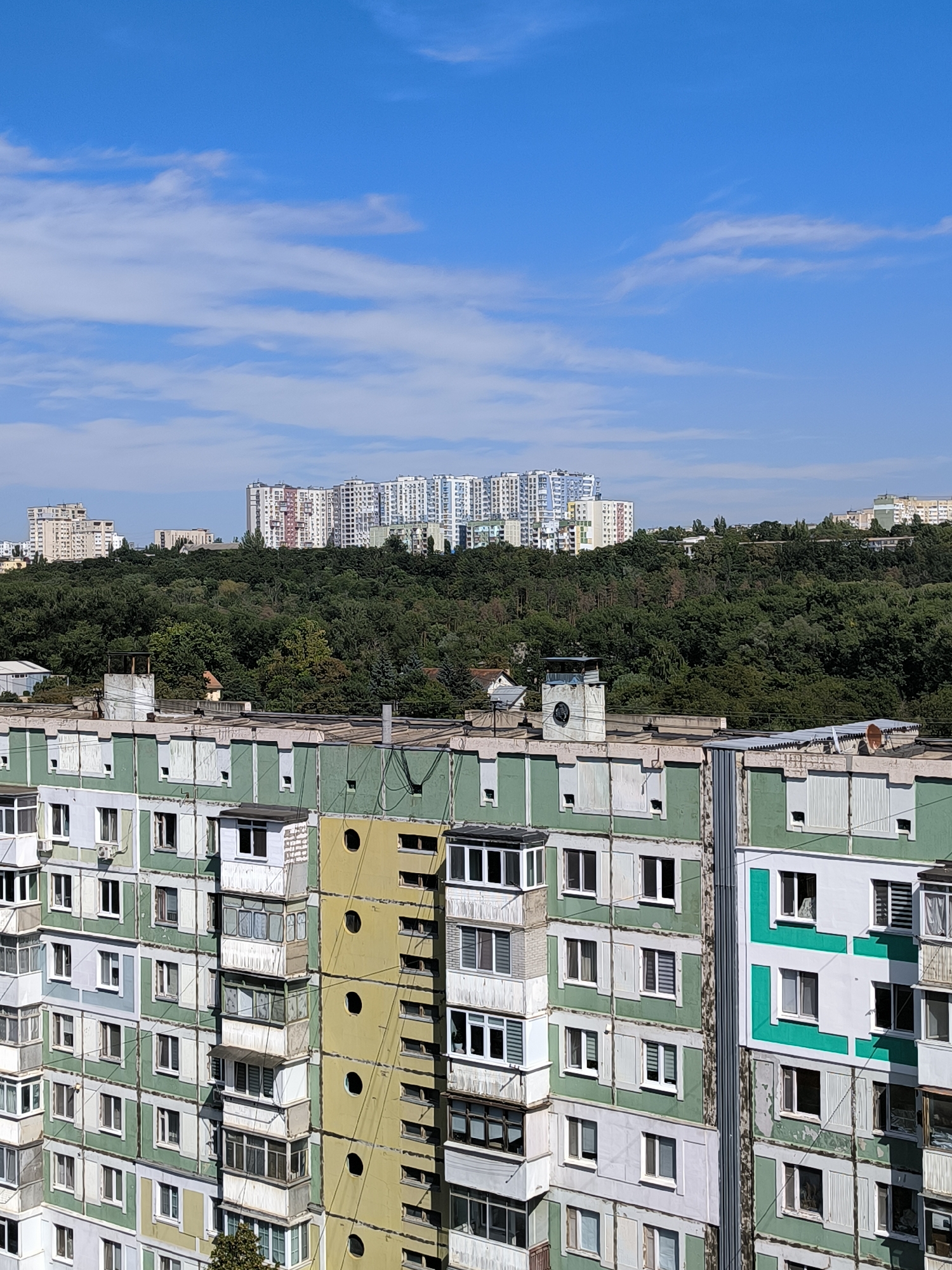 This is a high-angle, eye-level, exterior, full, daylight shot of a residential area in Chișinău, Moldova. The foreground is dominated by the facade of a multi-story apartment building painted in alternating sections of light green and white, with some sections also in yellow. The building features numerous windows, balconies, and air conditioning units. On the roof of the foreground building, there are vents and pipes. Behind the foreground building, a dense, dark green forest covers a hillside. Beyond the forest, a row of modern, multi-colored high-rise apartment buildings are visible under a clear blue sky with sparse white clouds. The overall impression is one of a dense urban landscape with a mix of older and newer residential architecture, surrounded by a significant green space.