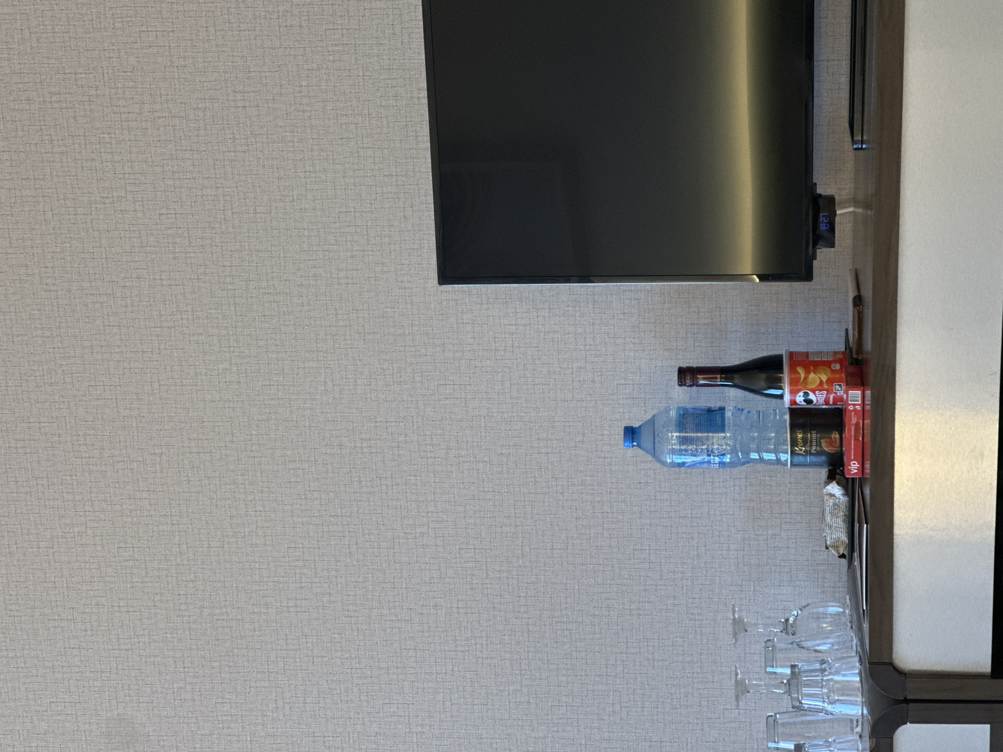 The scene is an interior shot of a room, potentially a hotel, featuring a television mounted on the wall above a built-in cabinet. The wall is covered in a neutral-colored, textured wallpaper. On the cabinet, which appears to be made of white and dark wood, there are several items: a bottle of wine, a bottle of water, a box of potato chips, a box of peanuts, several wine glasses, and a snack. The clock on the television reads 18:27. The location is possibly within the city of Plovdiv, Bulgaria.