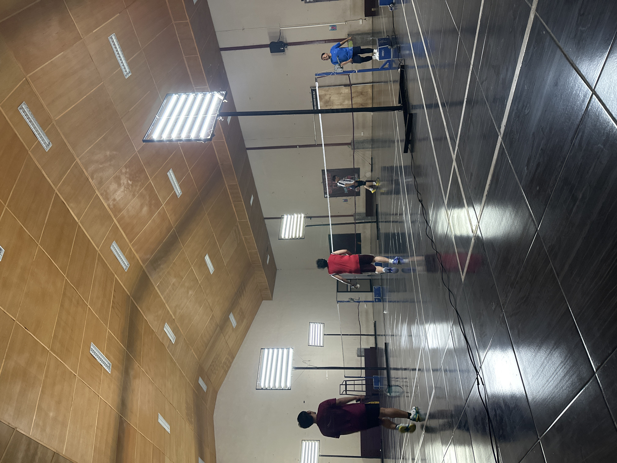 An indoor badminton court in the city of Batu, Indonesia, is depicted with four individuals engaged in a game. The court features a polished dark floor with white court lines, and a net divides the playing area. Overhead, the ceiling is constructed with wooden panels and illuminated by recessed fluorescent lights and two large, bright LED panel lights mounted on stands. The lighting creates strong reflections on the glossy floor.
On the left side of the court, a person wearing a maroon shirt and black shorts is seen from behind, holding a badminton racket. To their right, a person in a red t-shirt and dark shorts is in motion, facing away from the camera. Further across the net, a person in a striped black and white shirt and dark shorts is visible. On the far right, a person in a blue t-shirt and black shorts stands near the net. One shuttlecock is visible on the floor. The environment suggests a sports facility with an athletic atmosphere.