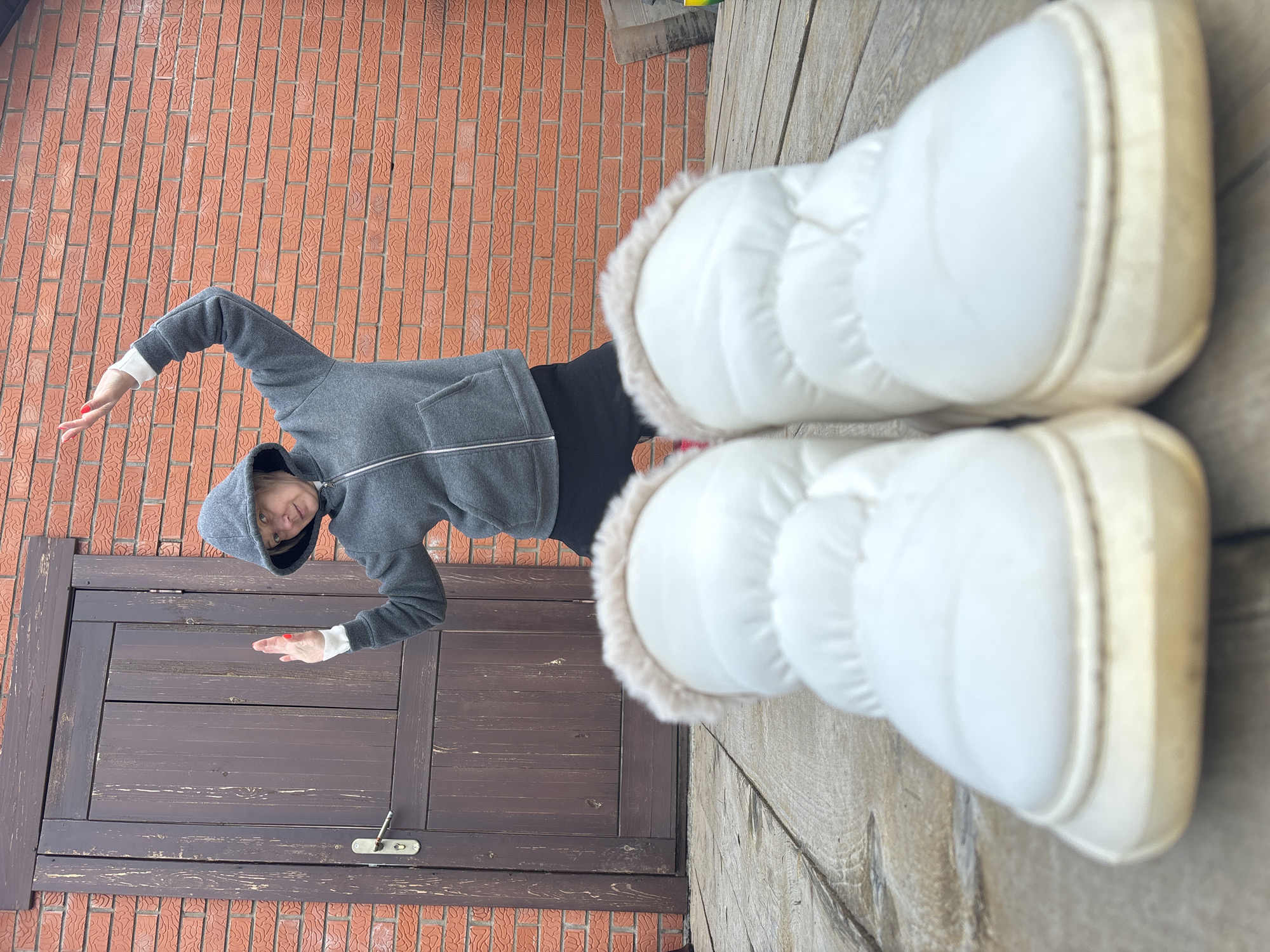 A woman is standing on a wooden deck in front of a brown wooden door and a brick wall. She is wearing a gray hooded sweatshirt and black pants, and has white snow boots with beige fur trim on her feet. Her hands are outstretched. The image may have been taken in Chaikovsky, Russia.