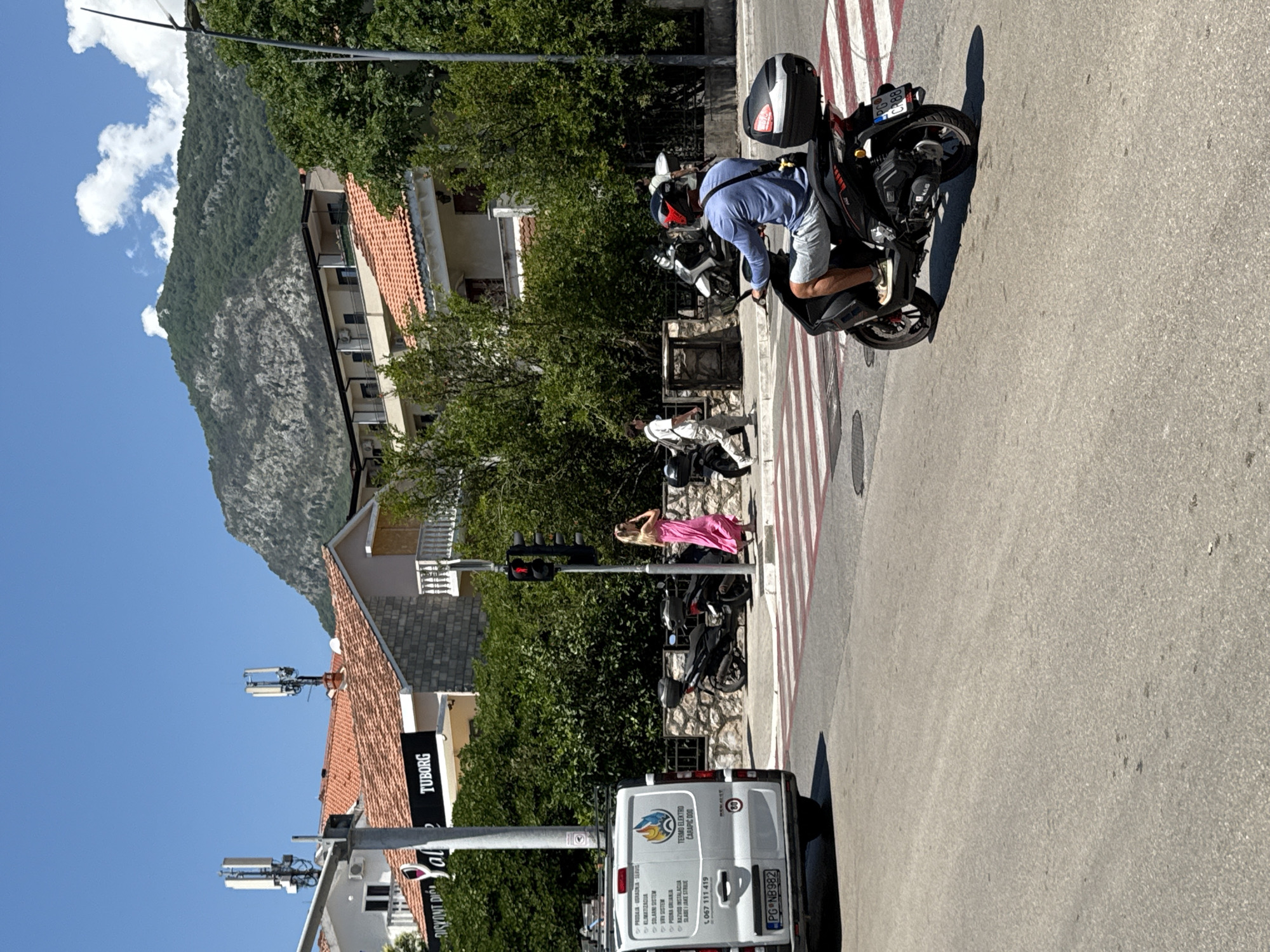 The image captures a street scene in Budva, Montenegro, on a bright, sunny day. A white van with branding for "TERMO ELEKTRO" is parked on the left, facing away from the camera. To the right, a pedestrian crossing is visible. A person in a pink dress is standing on the sidewalk, and another person in a white shirt and pants is walking away from the camera. A man wearing a blue long-sleeved shirt and grey shorts is riding a black scooter and approaching the pedestrian crossing. He is wearing a helmet with a red accent. In the background, buildings with red-tiled roofs and lush green trees are seen, with a large, tree-covered mountain forming the backdrop under a clear blue sky with some white clouds. Two telecommunication towers are mounted on top of a building on the left. A traffic light is visible, currently showing red.