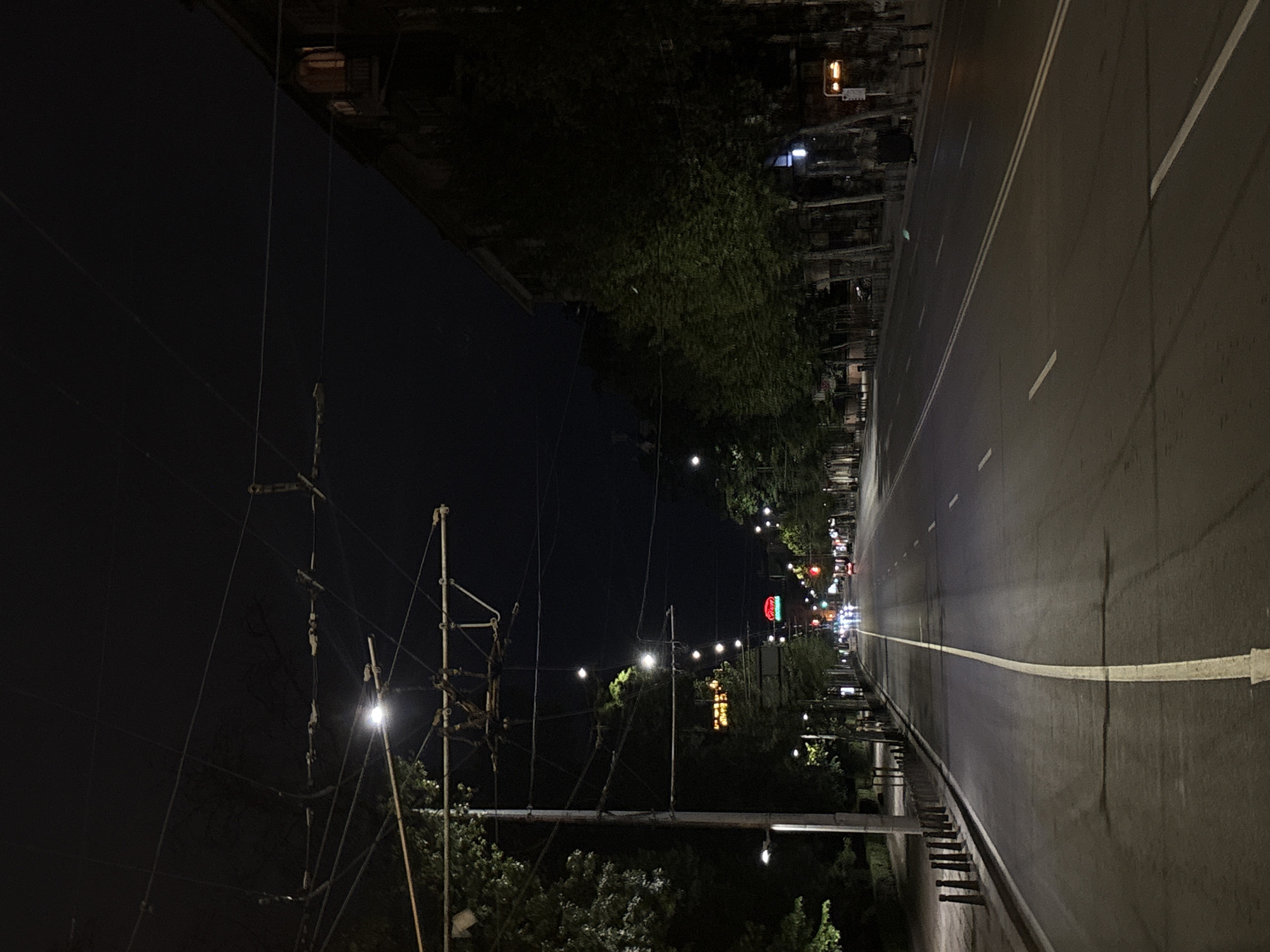 The image depicts a street scene in Sofia, Bulgaria at night. A road with two lanes marked by white lines stretches into the distance, lit by streetlights. The road is flanked by trees and buildings. A Coca-Cola sign is visible. A network of utility poles and wires dominates the left side of the frame against the dark sky. The lighting suggests an urban environment.