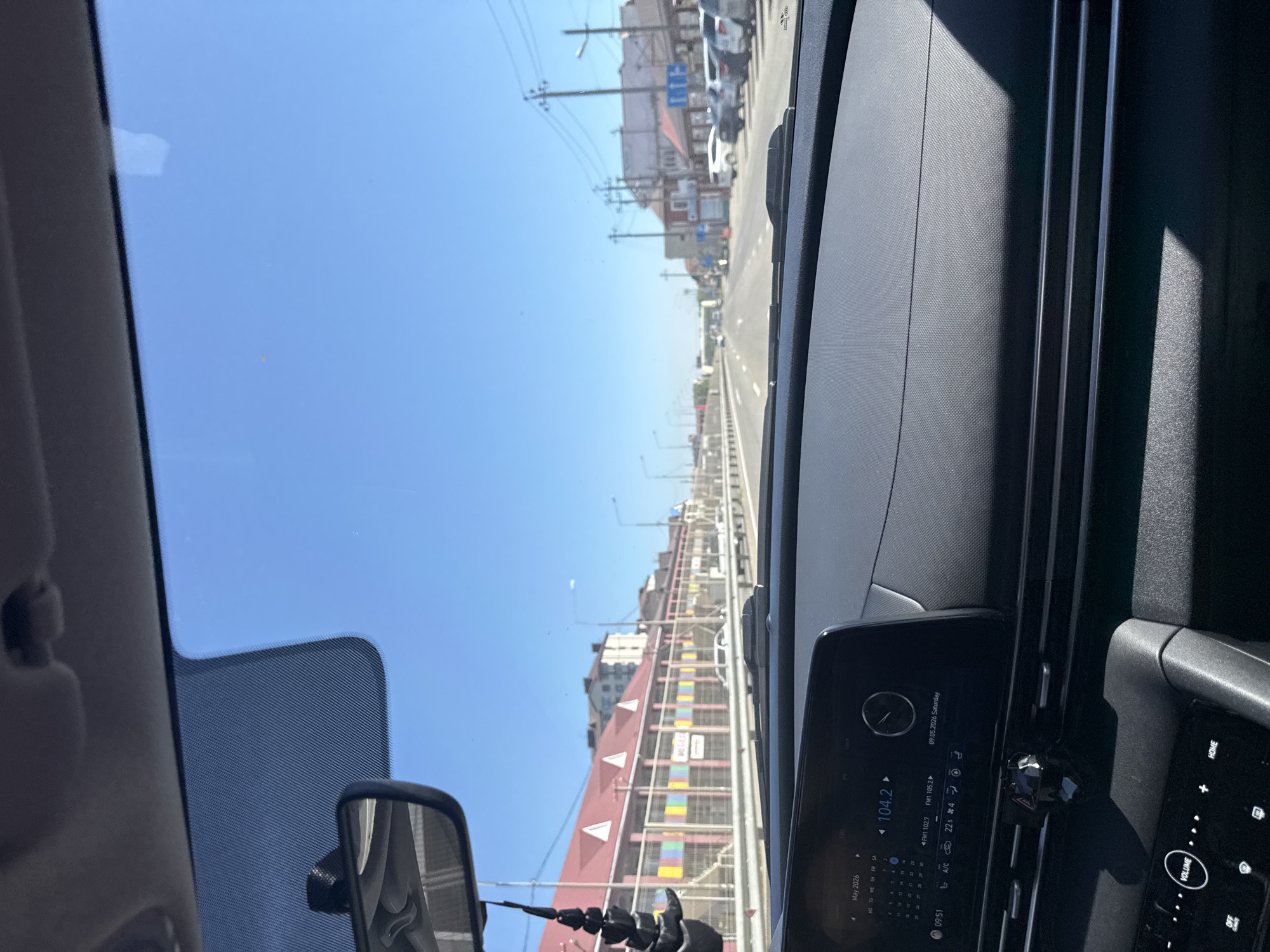 Captured from the interior of a vehicle, the image presents an upward-tilted view of a city street under a clear, blue sky. The street, lined with buildings and utility poles, stretches into the distance where vehicular traffic is visible. The camera's vantage point is from the dashboard of the car, where the infotainment system displays information including the date "09.05.2026 Saturday," a radio frequency "104.2," the interior temperature "22," and the month "May 2026." The steering wheel and other dashboard components are also visible, providing a perspective of the driver's point of view. A rearview mirror is present, adding depth to the scene. The environment is urban, with the focus on the street and its adjacent infrastructure.