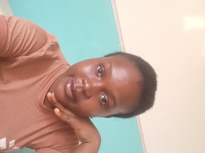 A forensic examination of the image reveals a medium close-up portrait of an adult female in an indoor setting within OGBOKO, Nigeria. The individual possesses dark skin, short, dark, closely-cropped hair, and directly faces the camera with a neutral expression. She wears a light brown, ribbed, short-sleeved top. Her left hand is raised, with fingers curled beneath her chin and thumb resting along her jawline. The background comprises a two-toned wall, featuring an upper section of light beige and a lower section of turquoise, with a horizontal demarcation line visible at shoulder level. Lighting is uniform, illuminating the subject's face. No specific interior identifiers are present to determine the precise location within Ogbolo beyond it being an interior space.