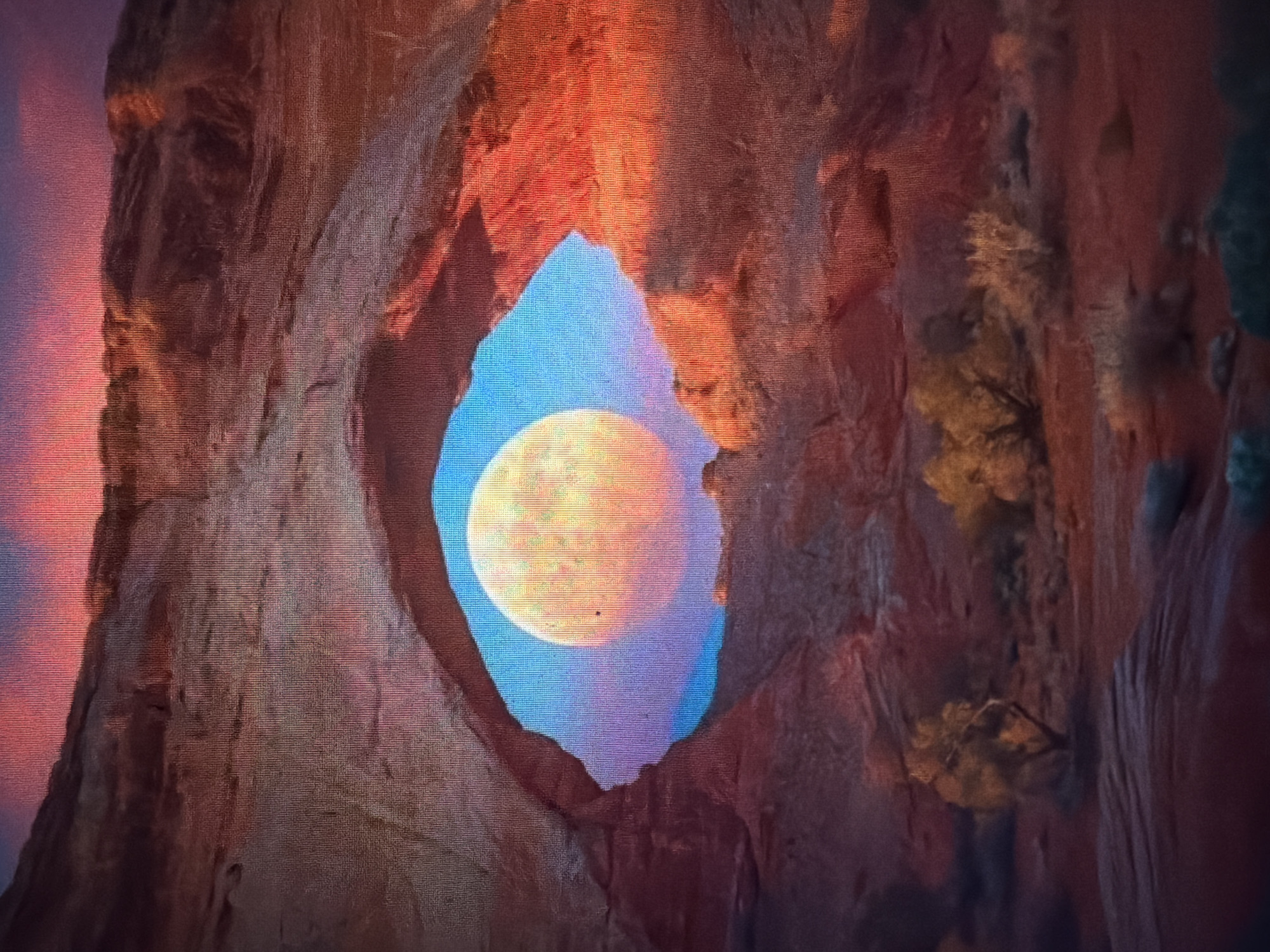 A full moon with a warm, orange hue hangs in a vibrant blue sky, framed by a natural rock arch. The rock formation is a deep reddish-brown, illuminated by the warm light of what appears to be sunset or sunrise. The sky above the arch transitions from a bright pink and orange near the horizon to a deep purple and blue at the top. In the foreground, silhouetted against the illuminated sky and rock, are sparse desert-like vegetation, including several small, leafless trees and shrubs. The ground is rocky and barren, with a reddish-brown hue. This scene depicts a natural landscape, likely a desert or canyon, with the moon prominently featured as the focal point. The image does not depict the city of Baan Mae Nam, Thailand.