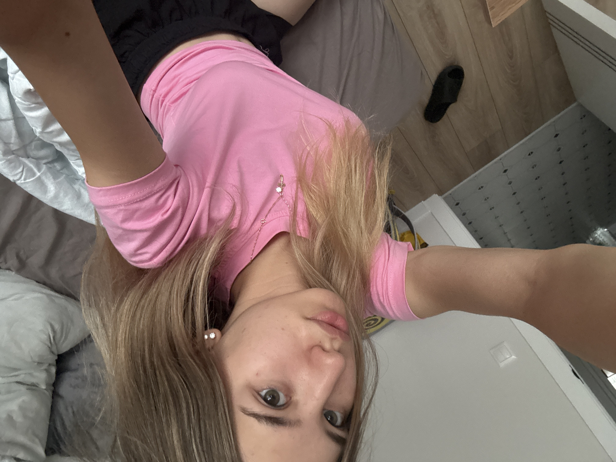 A young woman with blonde hair and green eyes is taking a selfie indoors. She is wearing a pink t-shirt and black shorts, and a silver necklace with a cross pendant. Her right arm is extended, holding the phone. In the background, a bed with grey and blue bedding is visible, as well as a tiled floor and wooden flooring with a black slipper. The overall setting appears to be a bedroom. There is no indication of the location being in Kazan, Russia.