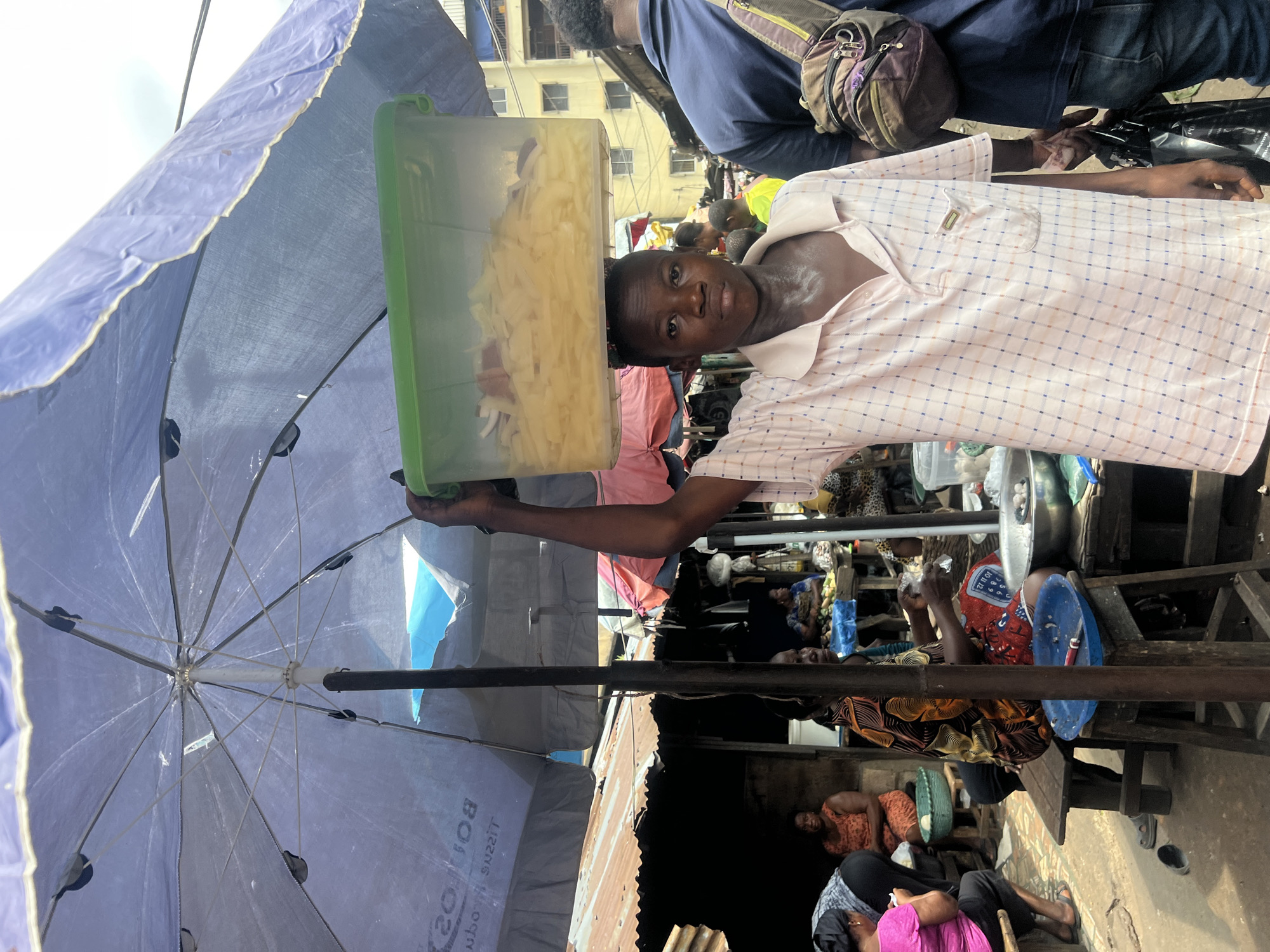 In Nkpor, Nigeria, a young boy, likely a vendor, is captured in a market scene. He stands under a large, blue umbrella, balancing a clear plastic container filled with sliced fruit on his head. The container is green and rectangular. The boy wears a light-colored, checkered polo shirt. Behind him, other market activities are visible, including stalls with tin roofs, other vendors, and shoppers. The ground is dirt and appears dusty. To the right, another person is partially visible, carrying a backpack and a black plastic bag. The overall impression is a bustling market environment.