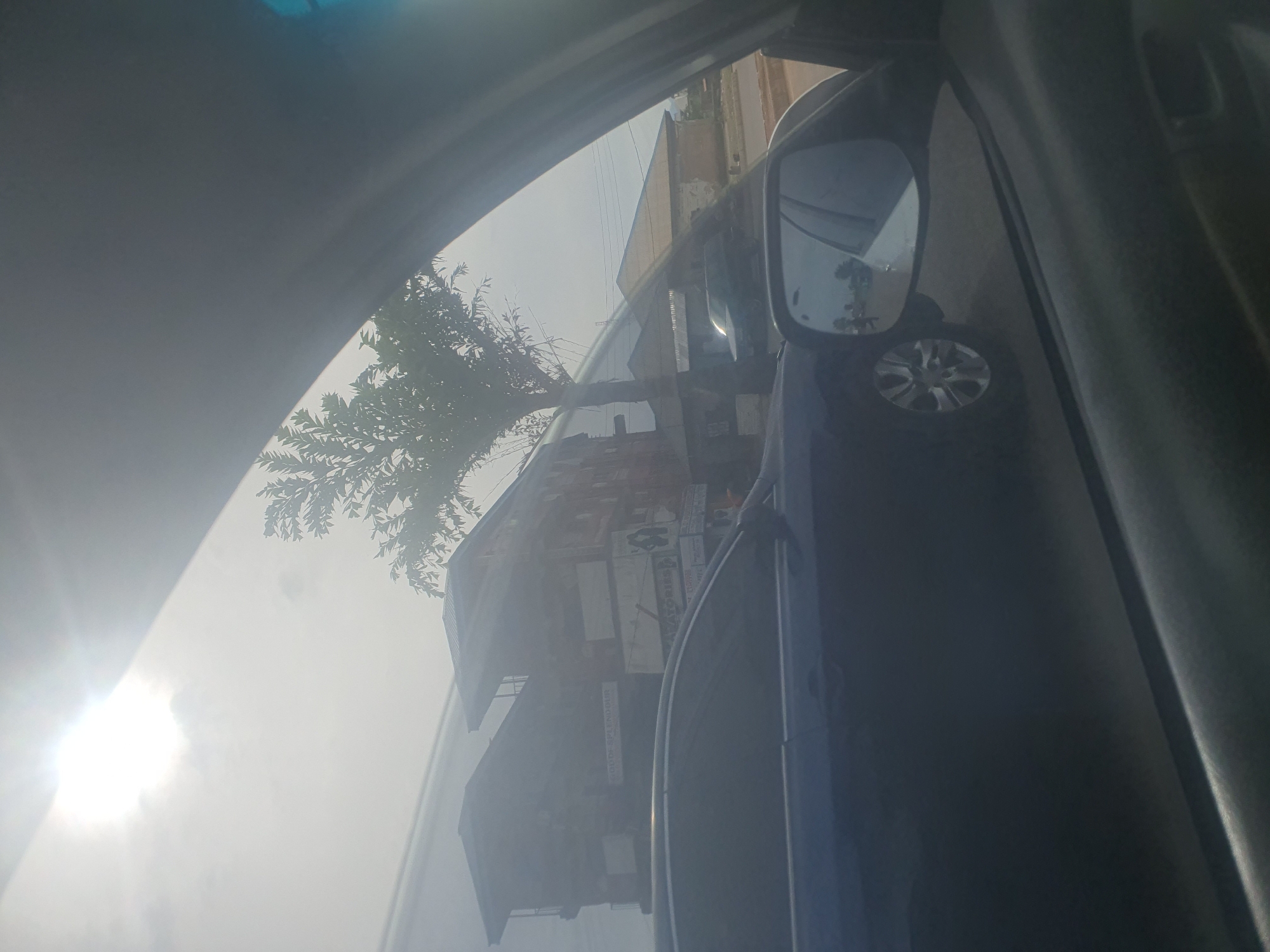 The image shows a view from inside a vehicle looking out at a street scene in Rafin Par, Nigeria. The sun is bright and partially obscured by clouds, casting a glare. In the foreground, a dark blue car is visible. Through the car's window and the side-view mirror, we see buildings, a tree, and other vehicles on the road. The buildings appear to be multi-story structures with signs, one of which reads "DE-SPLENDOUR" and another with text that is difficult to decipher. The overall impression is of a busy urban environment.