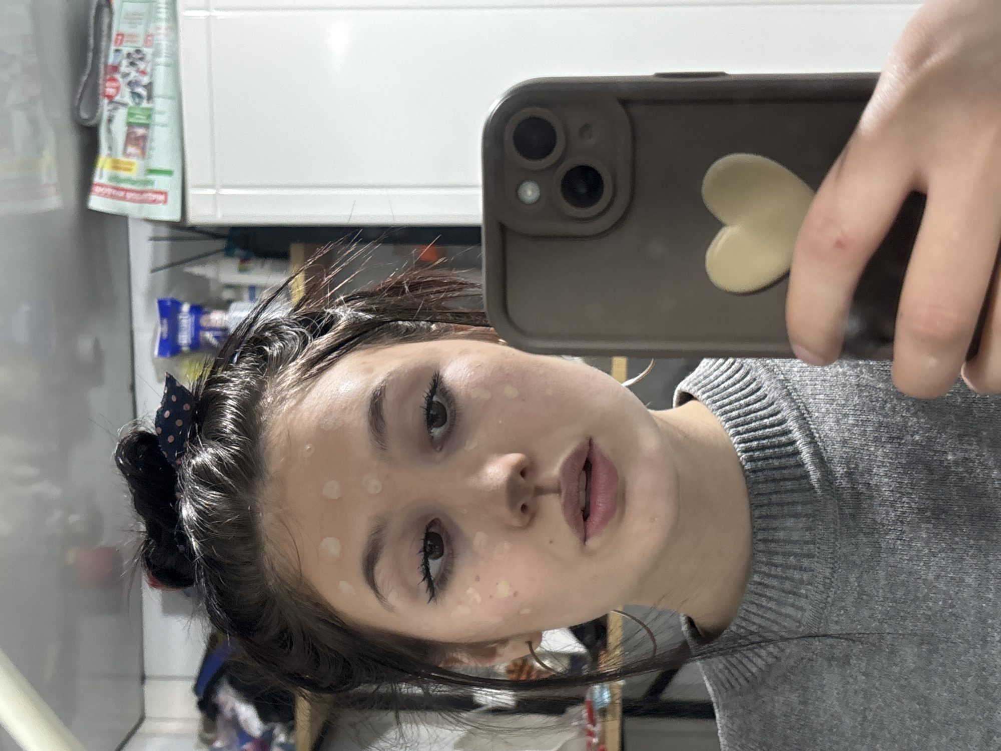 A young woman is captured in a mirror selfie, holding a dark-colored smartphone with her left hand. Her head is tilted, and she is looking directly at her reflection. She has dark hair, partially tied back with a dark polka-dotted scrunchie, with some strands framing her face. Her eyelashes are prominent with mascara, and her lips are slightly parted. Numerous small, light-colored dots, likely foundation or concealer, are visible across her forehead, cheeks, nose, and chin, indicating she is in the process of applying makeup or skincare.
She is wearing a gray knitted sweater with a ribbed collar. The smartphone case is dark, featuring a light-colored heart-shaped sticker on the back and two visible camera lenses with a flash/sensor. The background primarily consists of a white tiled wall. In the upper left corner of the image, reflected in the mirror, a blurred item resembling a shopping bag or flyer hangs, displaying partial Russian text which includes "РЯДОМ С ДОМОМ" (Near the house) and "АПРЕЛЬ" (April), along with some prices. The scene appears to be an indoor setting, likely a bathroom or vanity area.