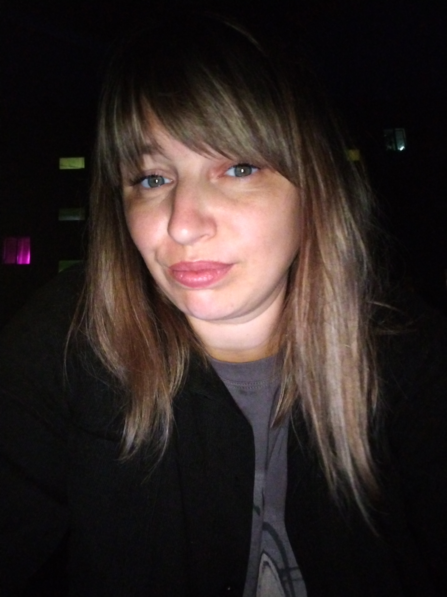 The image is a selfie of a woman with light brown hair and blue-green eyes. She is wearing a black jacket over a grey t-shirt. Her lips are painted pink. In the background, there are some blurred lights, one of which appears to be purple. The image is dark, suggesting it was taken at night. This scene could be anywhere as there are no specific details to identify the location.