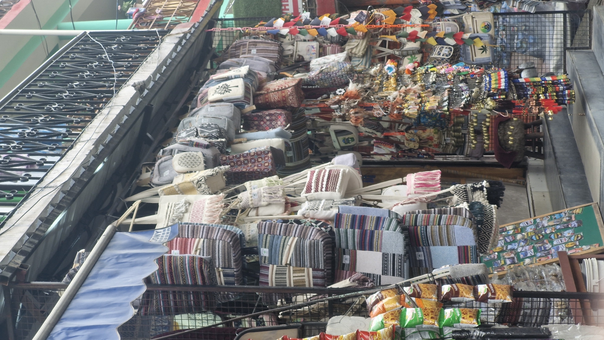 The image depicts a street market stall in Vashisht, India, likely a tourist area, displaying a variety of handcrafted bags and souvenirs. The stall is a colorful and cluttered arrangement of textile goods. Numerous woven bags, predominantly in earthy tones and featuring geometric patterns, are hung from the awning and the stall's structure. Some bags are adorned with tassels, fringes, and embroidery. Mixed in with the bags are small decorative items, possibly keychains or ornaments, made of colorful threads and beads, hanging in dense clusters.
Towards the left side of the frame, bags are stacked vertically on a rack, with a display of packaged snacks and bottled items visible behind them. A green display board featuring numerous small, colorful stickers or magnets is propped up near the front. Further to the right, a small, furry stool or pouf with a dark red covering is placed on the steps leading up to the stall. The backdrop shows a building with a green facade and a decorative metal railing on an upper level. The scene is illuminated by daylight, suggesting it is an outdoor market. No people are clearly visible in the image, but the presence of the merchandise indicates a commercial setting.