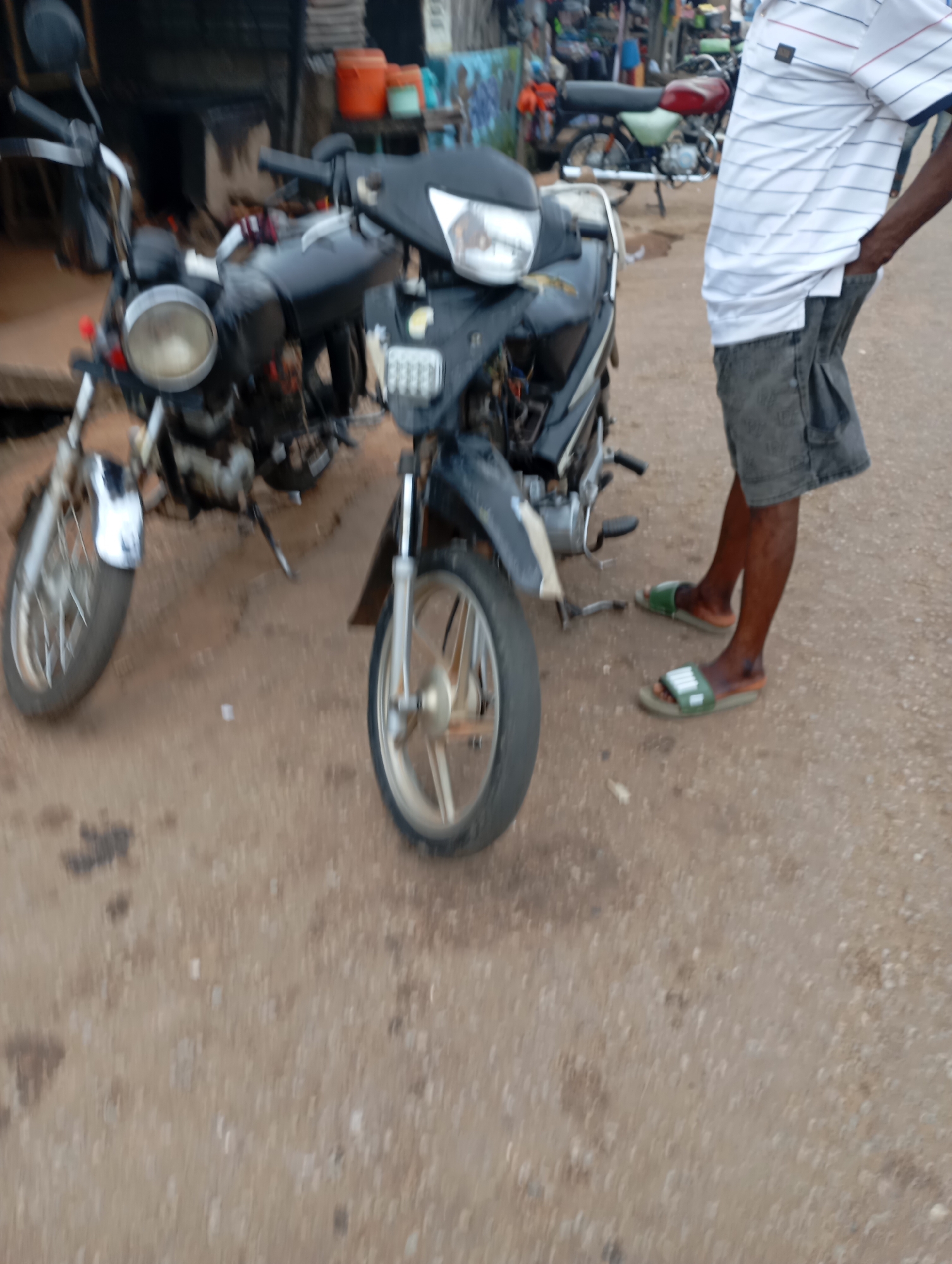 In Shendam, Nigeria, a low-angle, slightly blurred outdoor shot captures two motorcycles parked on a dirt road. The motorcycle on the left is predominantly black with a chrome headlight and a silver front fender. The motorcycle on the right, also black, has a more angular front fairing and a silver exhaust pipe visible. A man stands to the right of the motorcycles, his body angled away from the camera. He is wearing a white polo shirt with blue stripes and dark, possibly grey, cargo shorts. He is also wearing green slide sandals. The environment appears to be a roadside setting, with a building or structure visible in the background to the left, featuring orange and blue containers on a shelf. The ground is a mix of reddish-brown dirt and small stones.